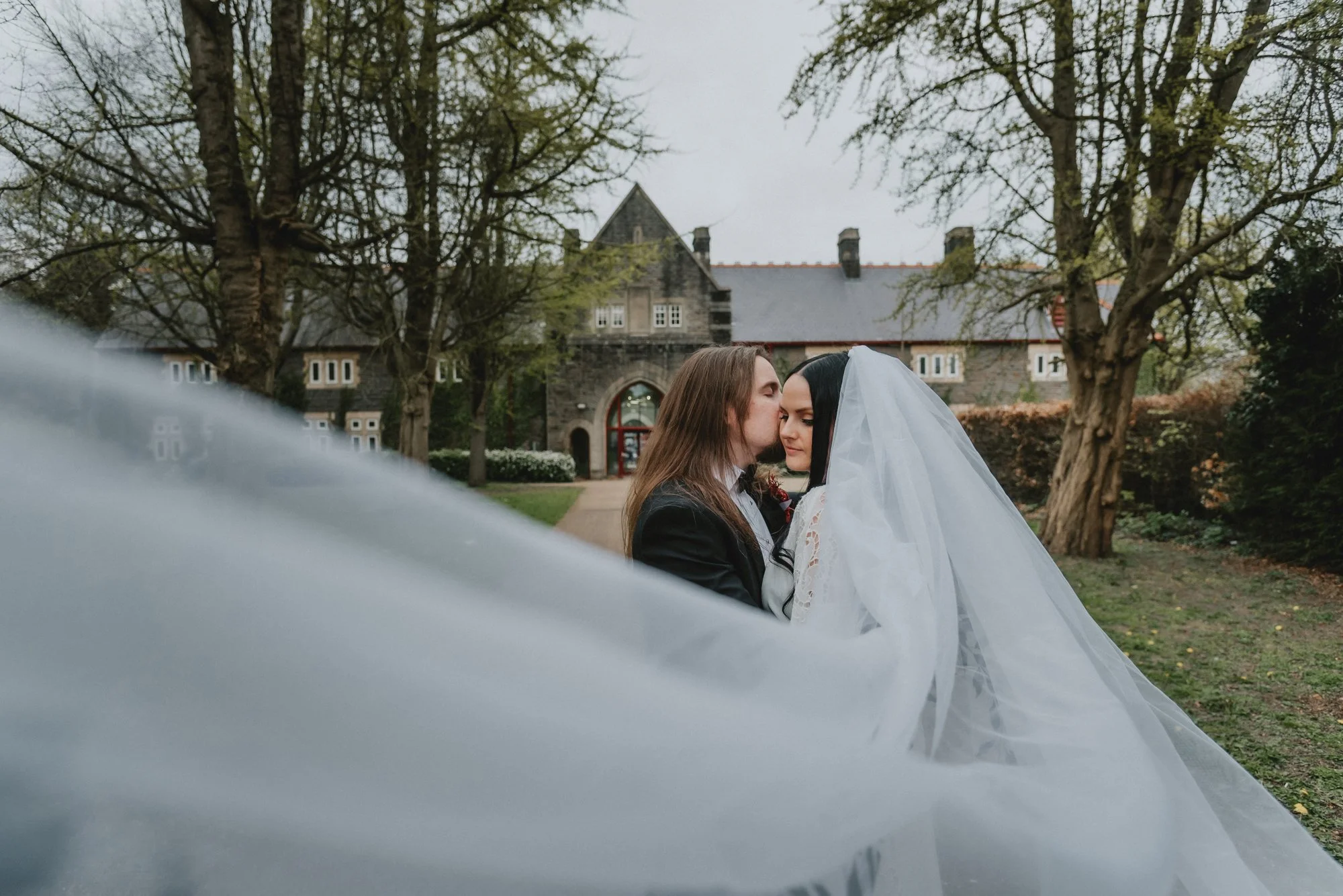 A bride and groom standing close together outdoors on a cloudy day, with the bride wearing a white veil and lace dress, and the groom in a black suit. There are trees and a stone building in the background.