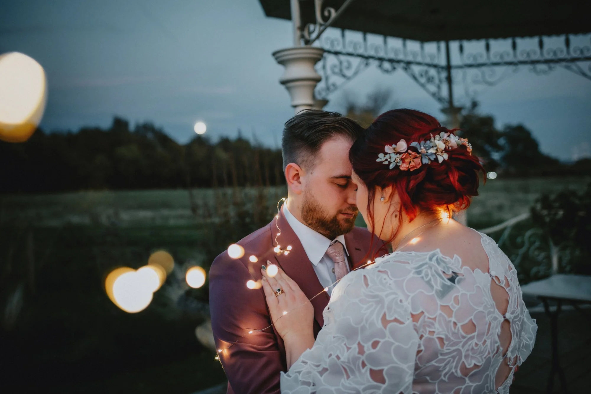 A bride and groom embrace warmly during their outdoor wedding at dusk, with string lights wrapped around them, under a decorative gazebo in a lush natural setting.