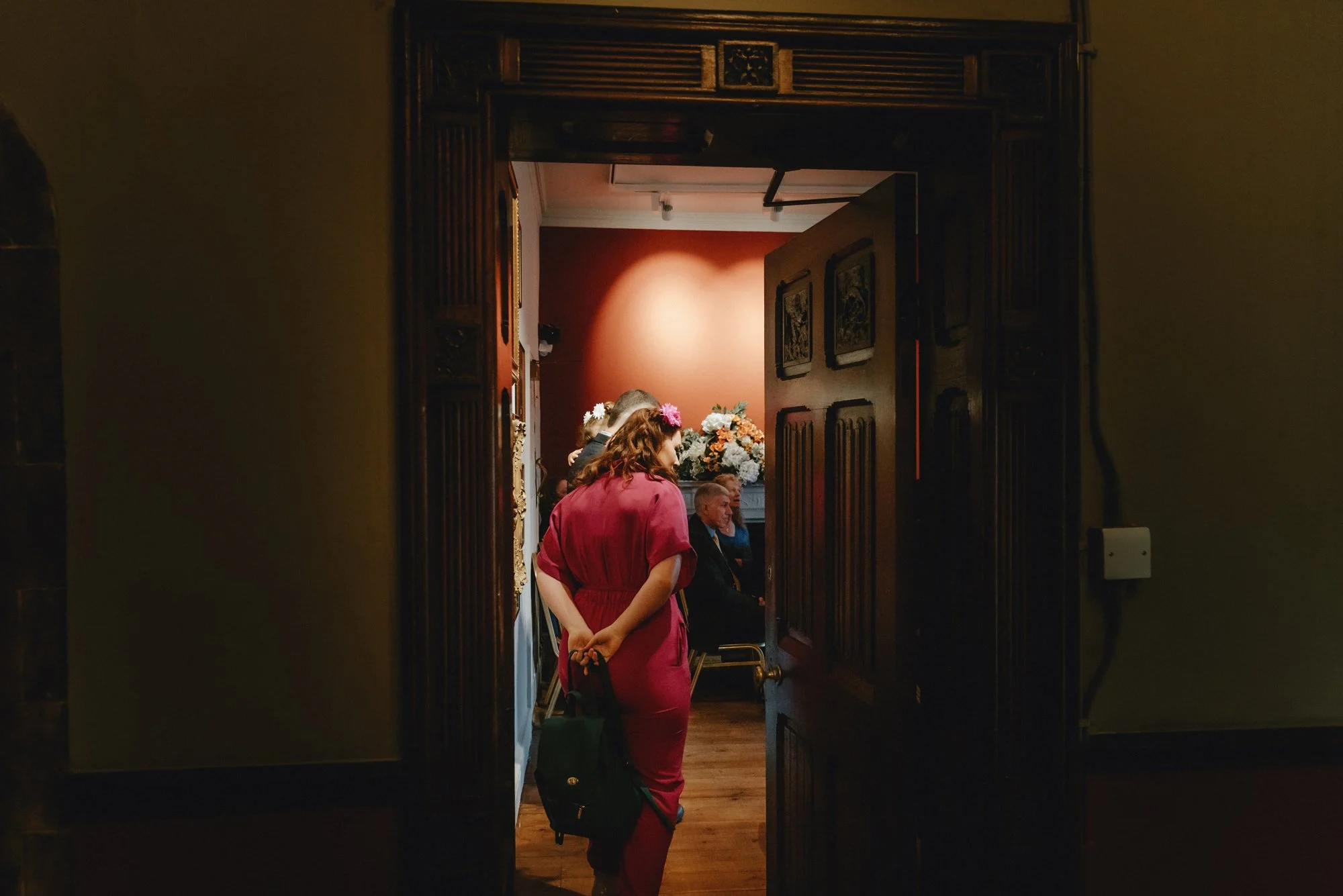 People gathered in a room with floral decorations and wood-paneled walls, seen through an open doorway.
