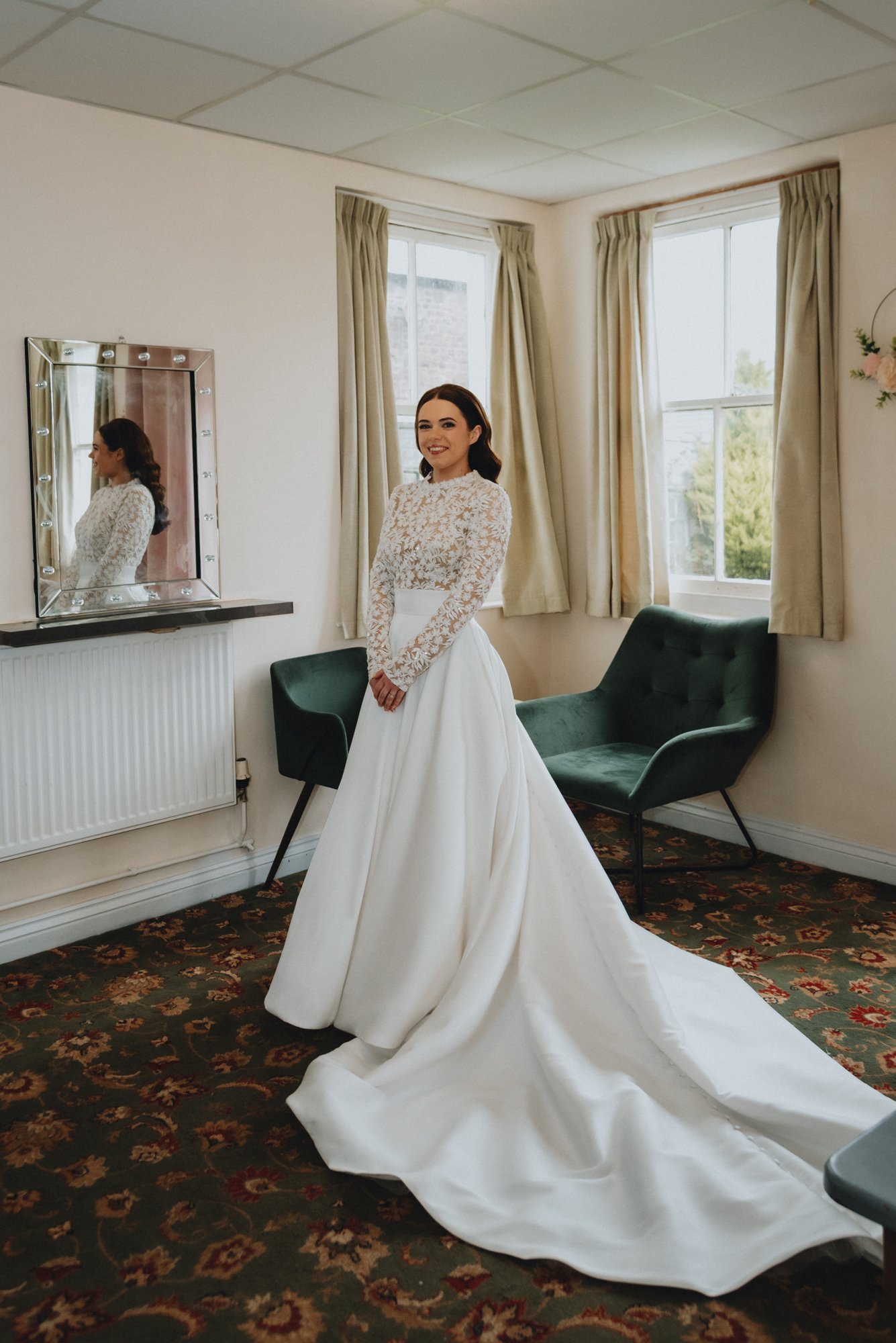 A woman in a white wedding gown standing in a room with natural light, a large window with beige curtains, a green armchair, a mirror with lights, and a floral carpet.