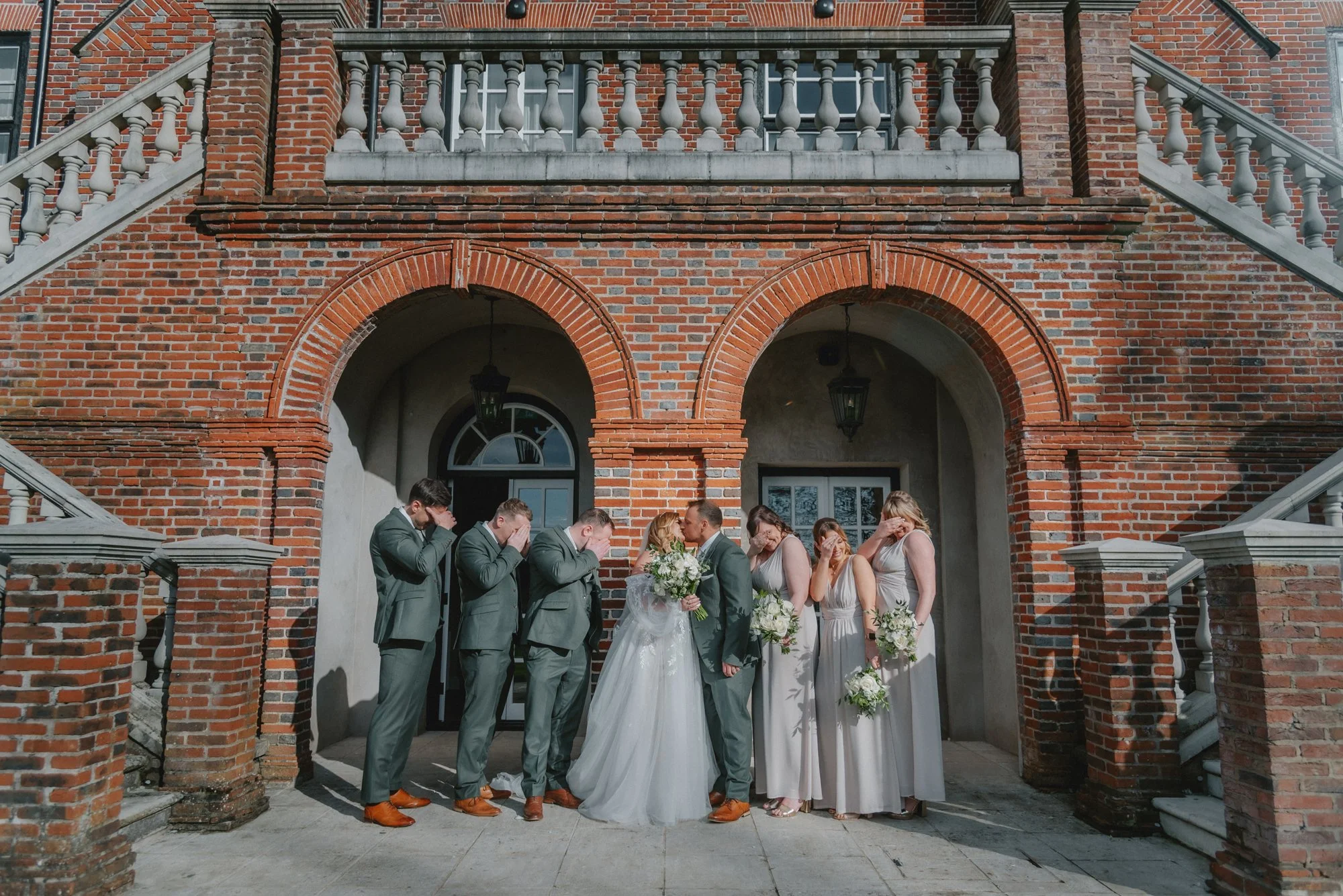 A wedding party stands outside a brick building with arched doorways. The bride and groom are in the center, kissing, with the bride holding a bouquet. Bridesmaids and groomsmen stand on either side, some covering their eyes.
