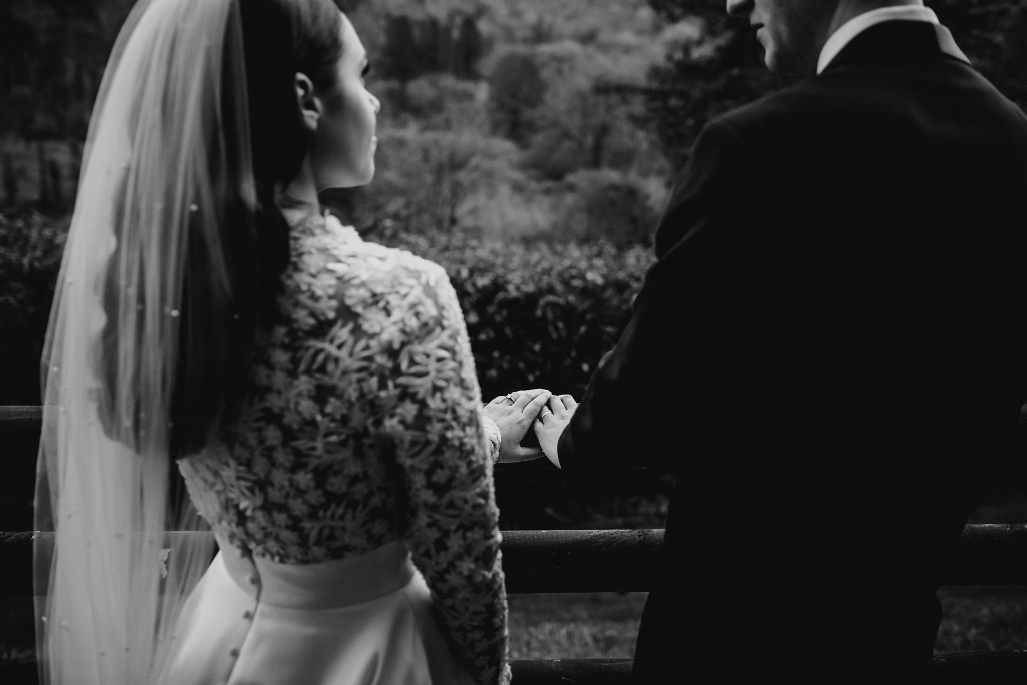 Black and white photo of a bride and groom holding hands while standing on a bridge outdoors, facing each other, with trees in the background.