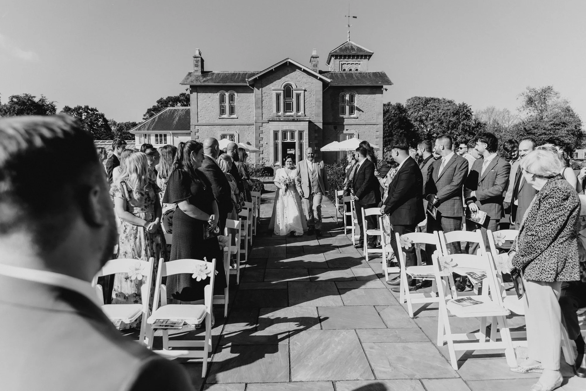 A black-and-white photo of a wedding ceremony outdoors, with guests seated on white chairs arranged on either side of an aisle, leading to a bride and groom walking down the aisle towards a large house or mansion in the background.