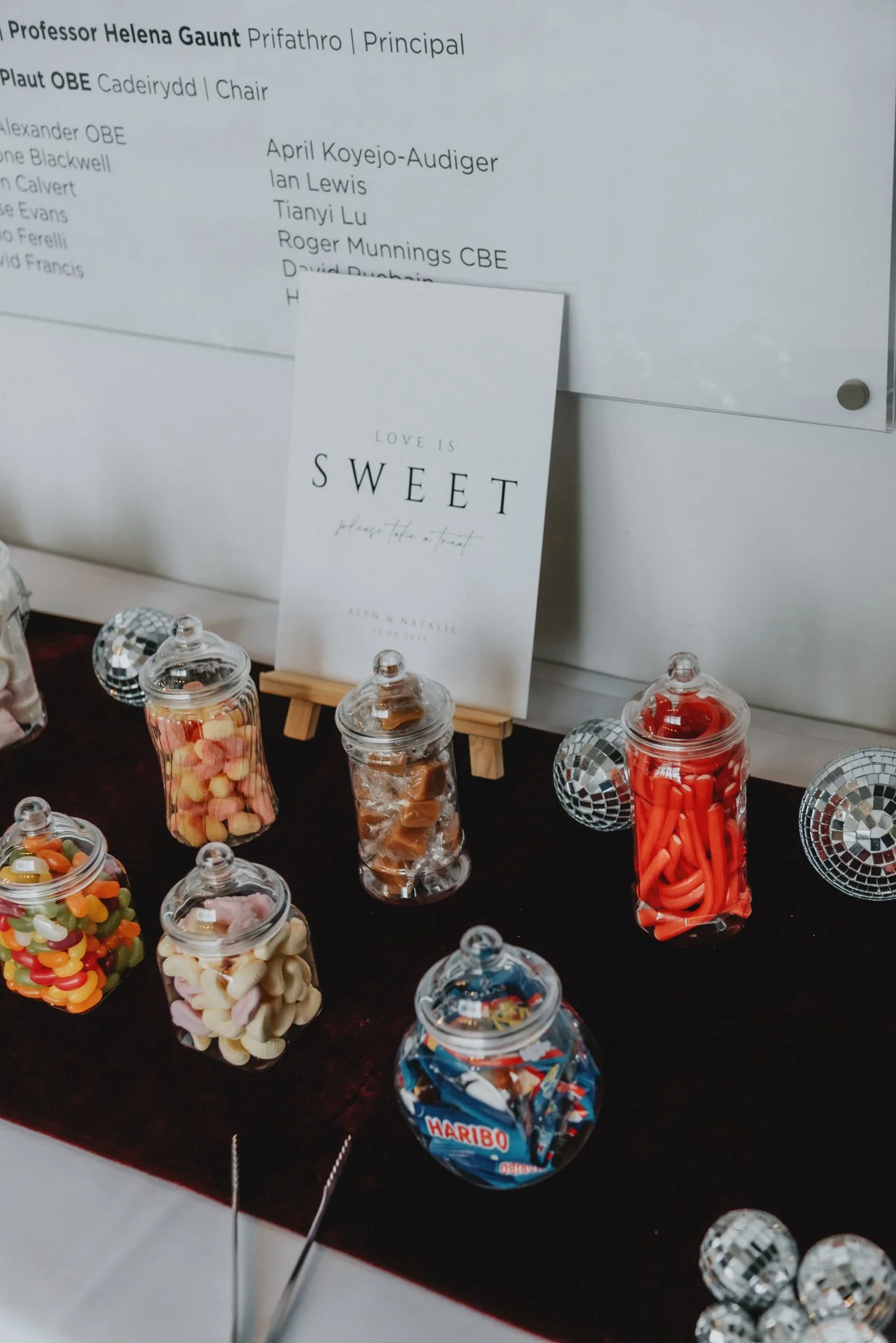 Jars of colorful candies, including gummy bears, licorice, and Haribo gummies, arranged on a table with a dark surface, decorated with small disco ball ornaments, and a white sign in the background with wedding details and a card that says "Love is S
