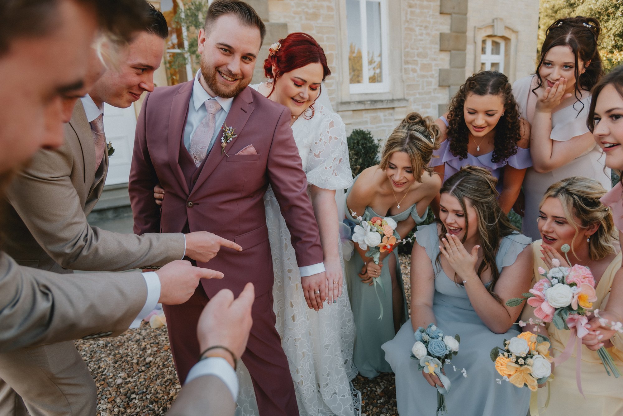 Group of smiling wedding guests looking at something amusing, gathered outdoors in front of a stone building, with women holding bouquets of flowers.