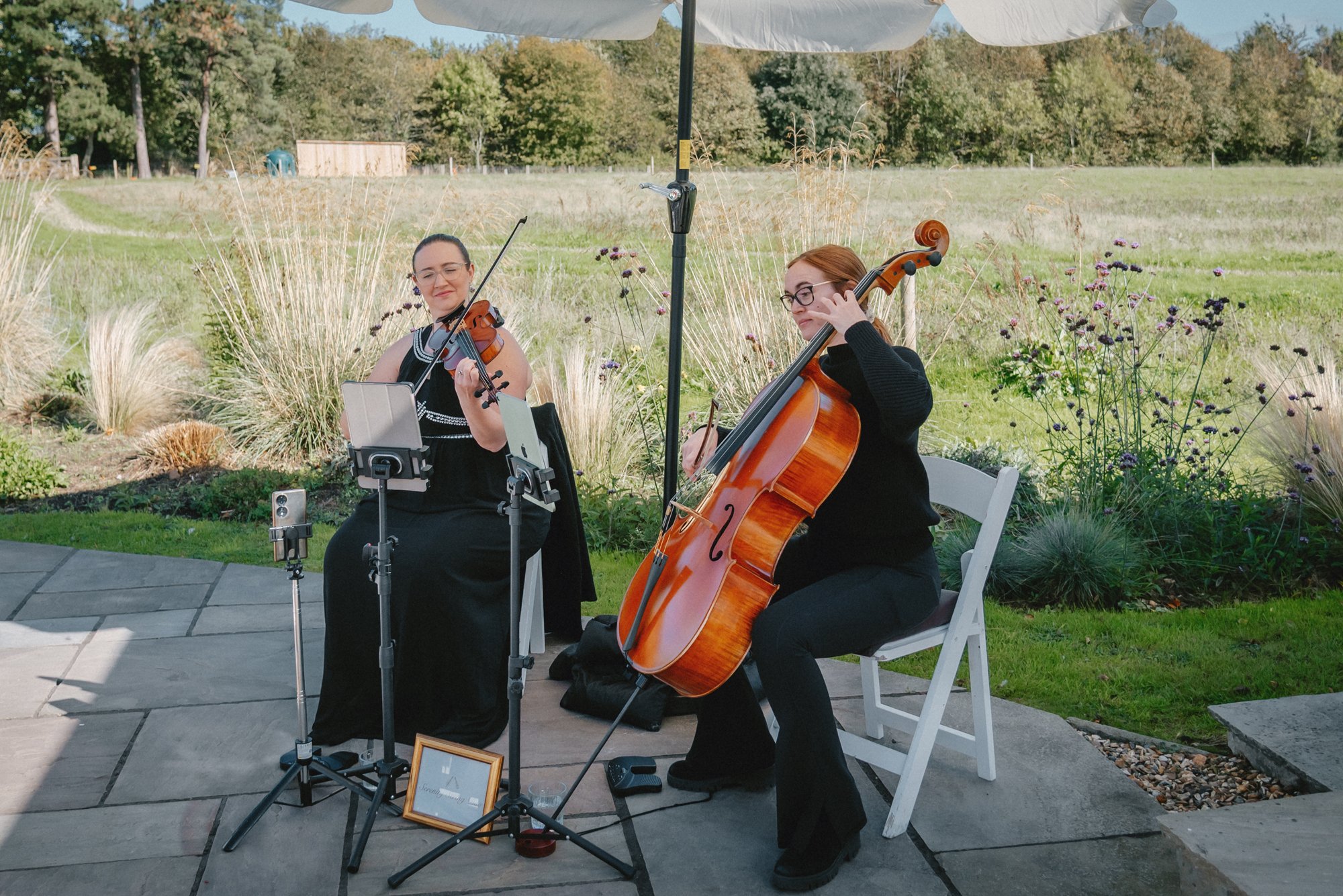 Two women playing string instruments, a violin and a cello, outdoors under a large white umbrella on a patio with a grassy field and trees in the background.