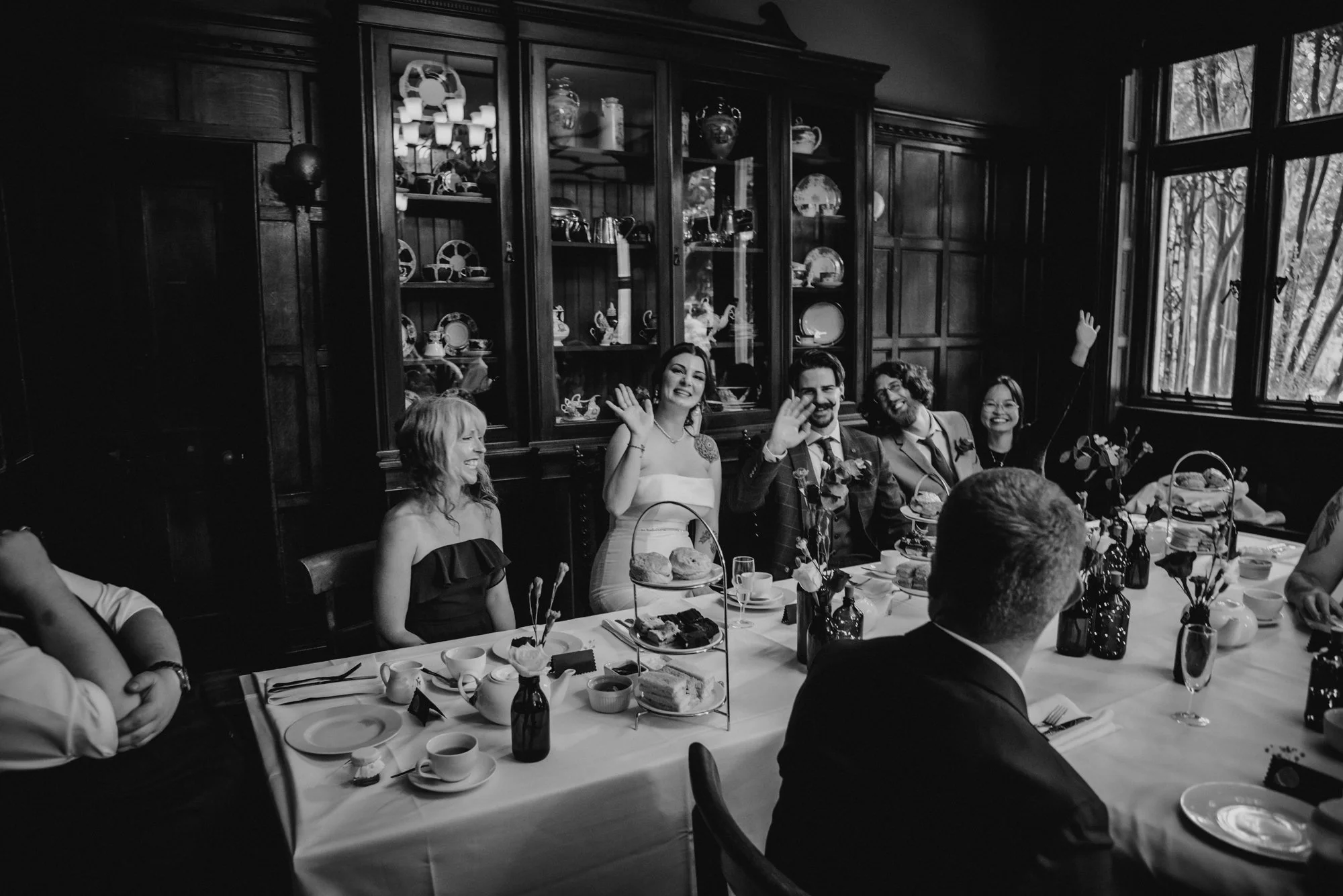 Black and white photo of a group of people at a dinner table, with some smiling and waving at the camera, in a room with wooden paneled walls and china displayed in a cabinet.