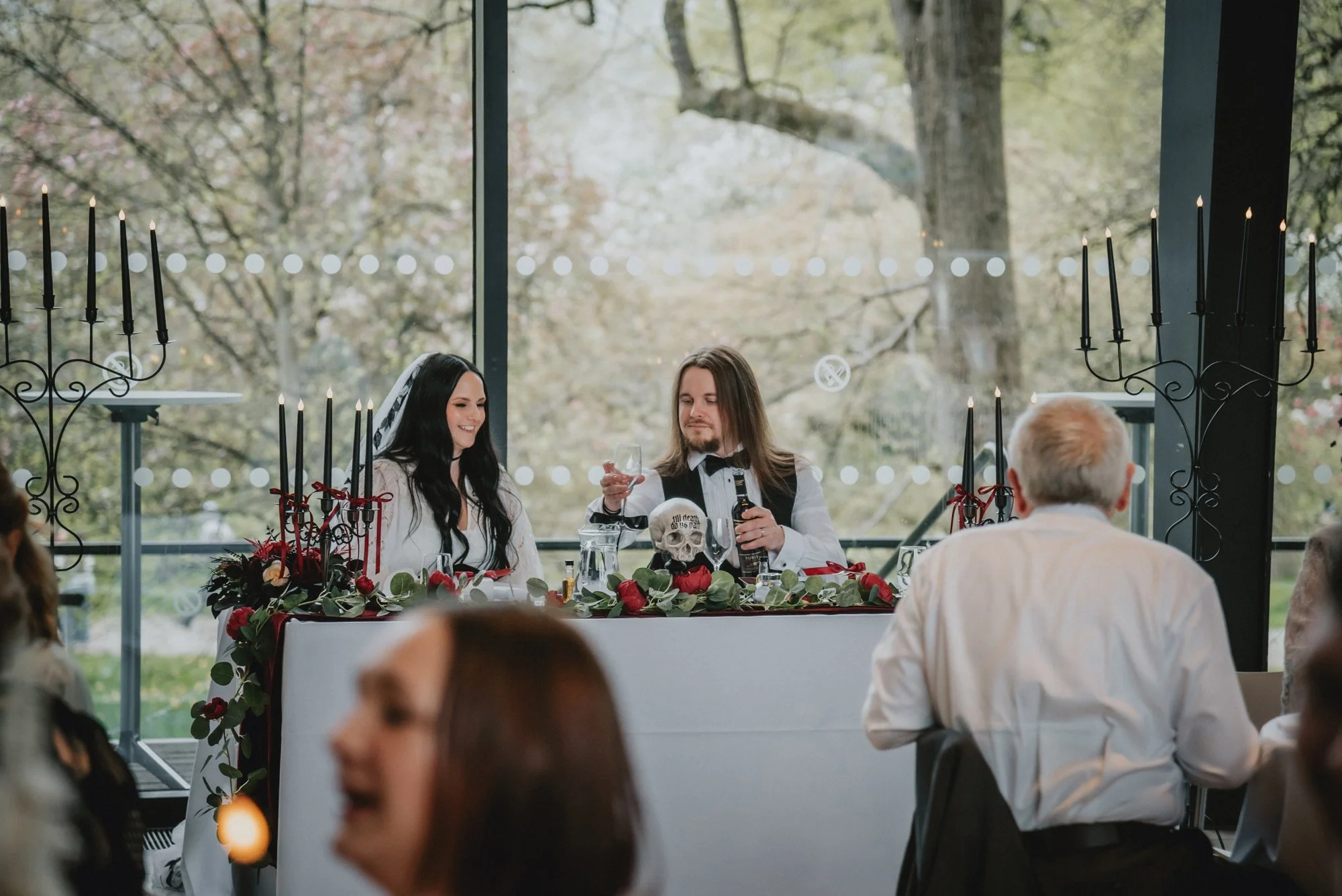 A wedding reception with a bride and groom dressed in formal attire, sitting at a decorated table inside a venue with large windows and outdoor greenery in the background. The table features a floral arrangement with red and green decorations and bla