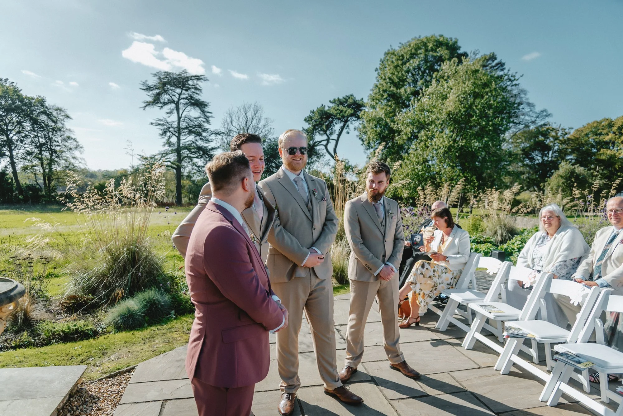 Groom and groomsmen standing in outdoor wedding ceremony with guests seated on white chairs, surrounded by trees and greenery on a sunny day.