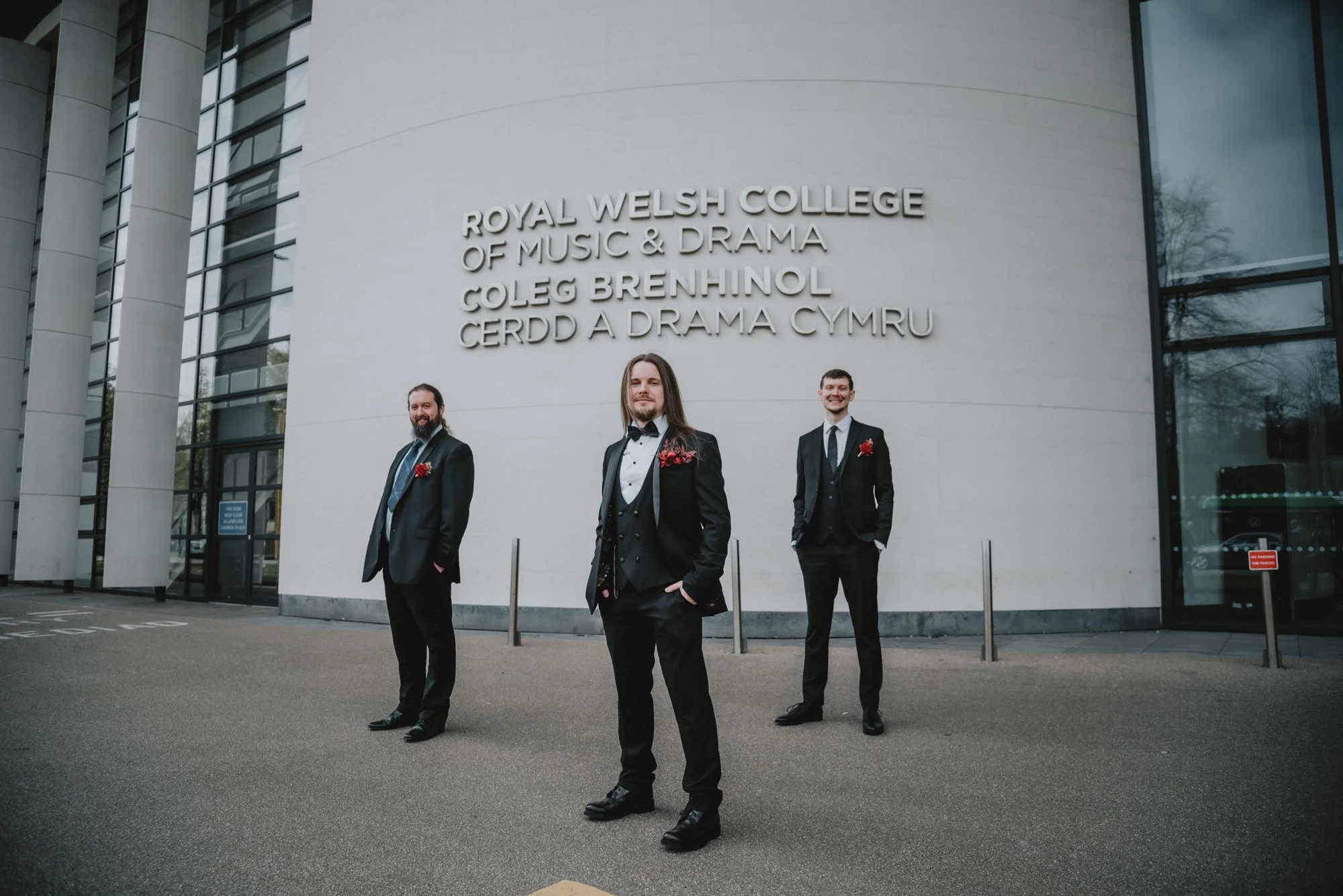 Three men in formal suits standing outside the Royal Welsh College of Music & Drama building, with the college's name on the wall behind them, one in the center and two on either side, all smiling.