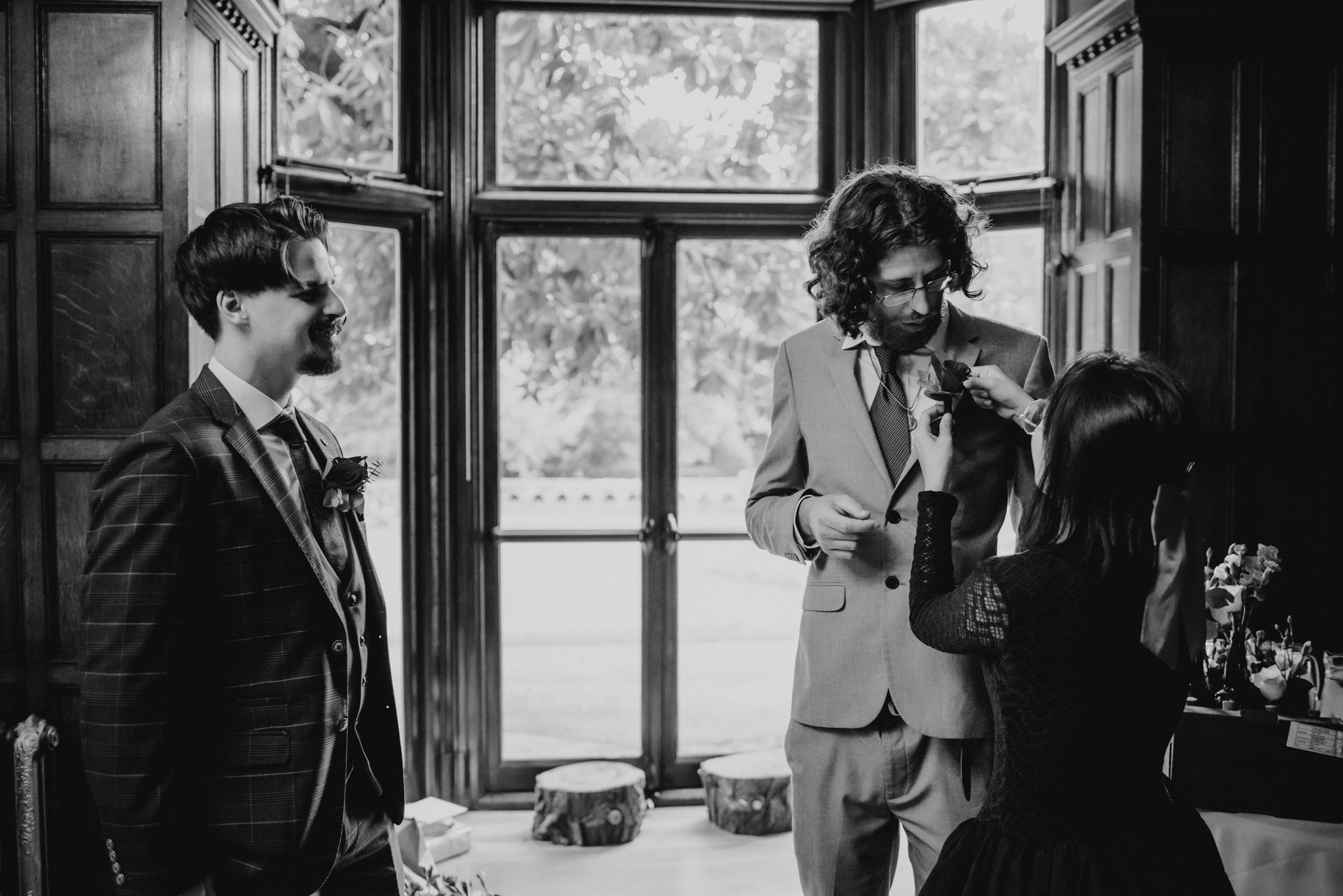 A Black and white photo of three people inside a wooden room with large windows. One man is dressed in a checked suit, standing on the left, smiling and looking at the other two. The man on the right, wearing glasses and a light suit, is pinning a bo