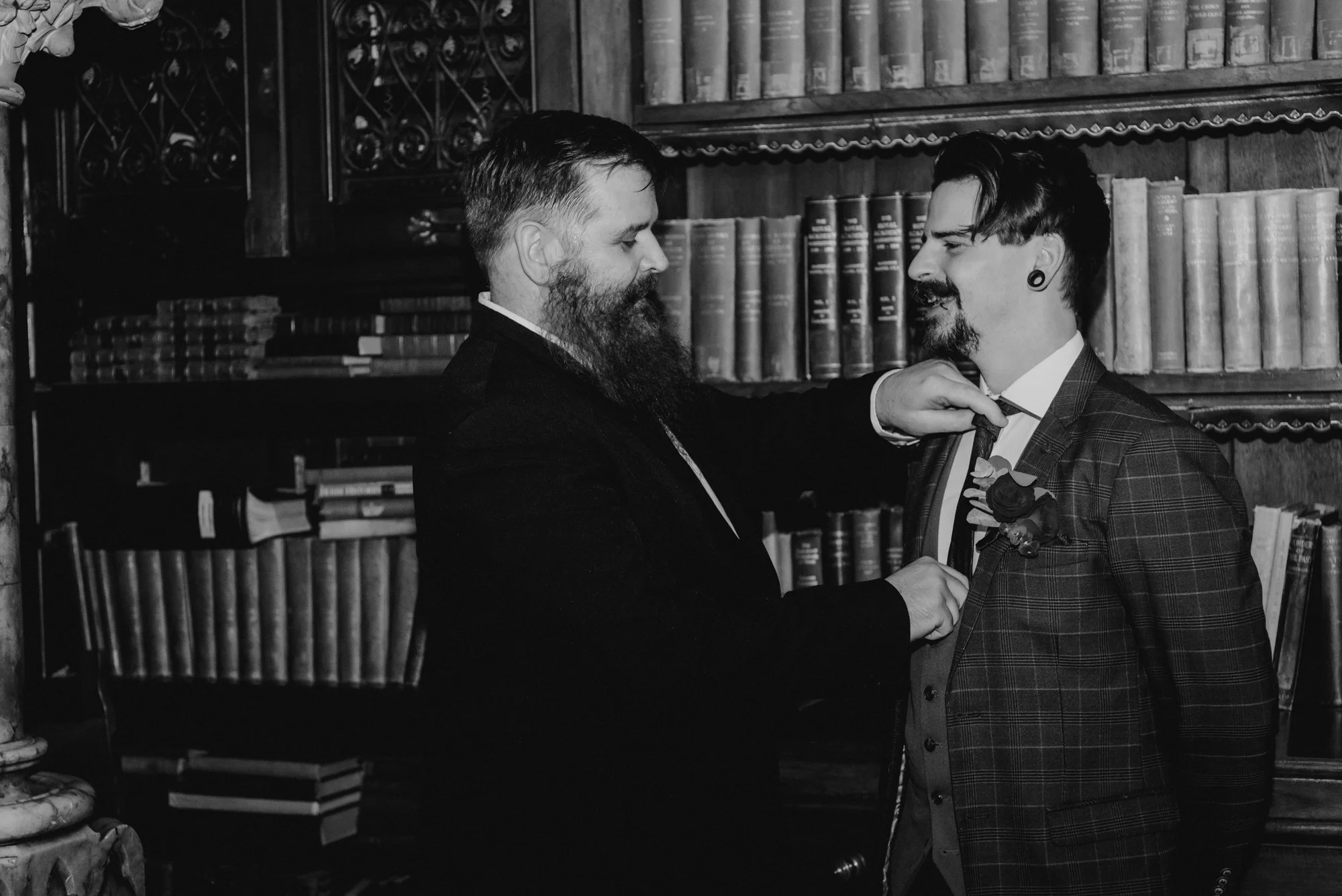 A black and white photo of two men in a library, with one man adjusting the other's tie. The man on the left has a beard and is wearing a suit, and the man on the right has a mustache and is wearing a plaid blazer with a boutonniere.