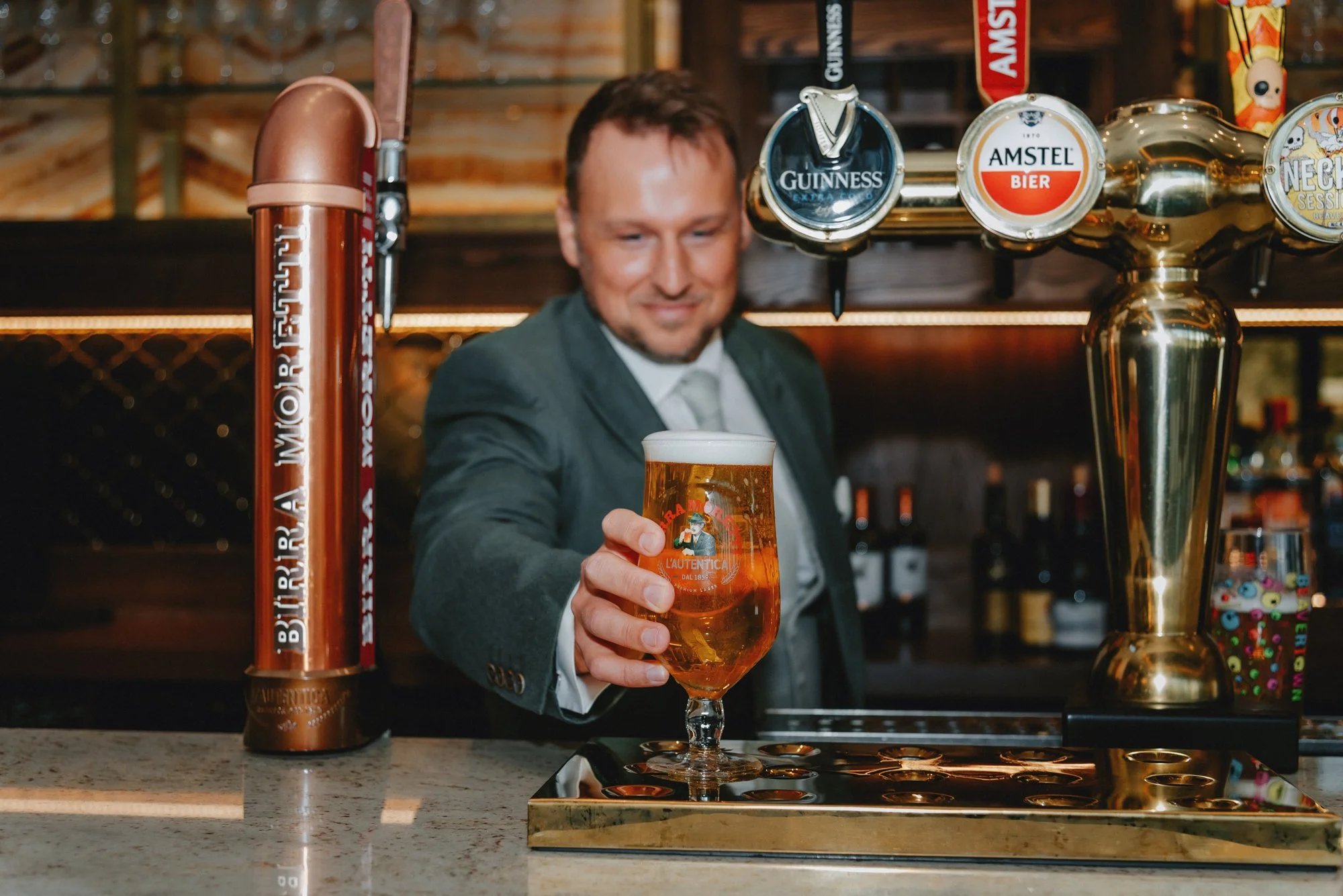 A bartender in a suit pouring a glass of beer in a bar with beer taps in the background.