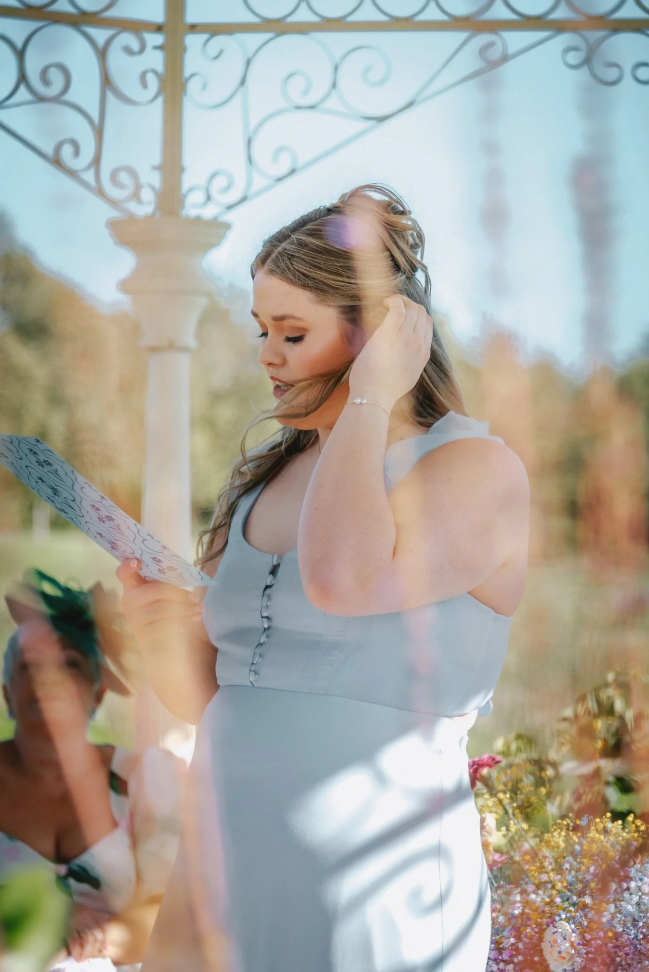 A young woman in a light blue dress with a button-down front, looking at a card, standing outdoors with trees and floral arrangements in the background.