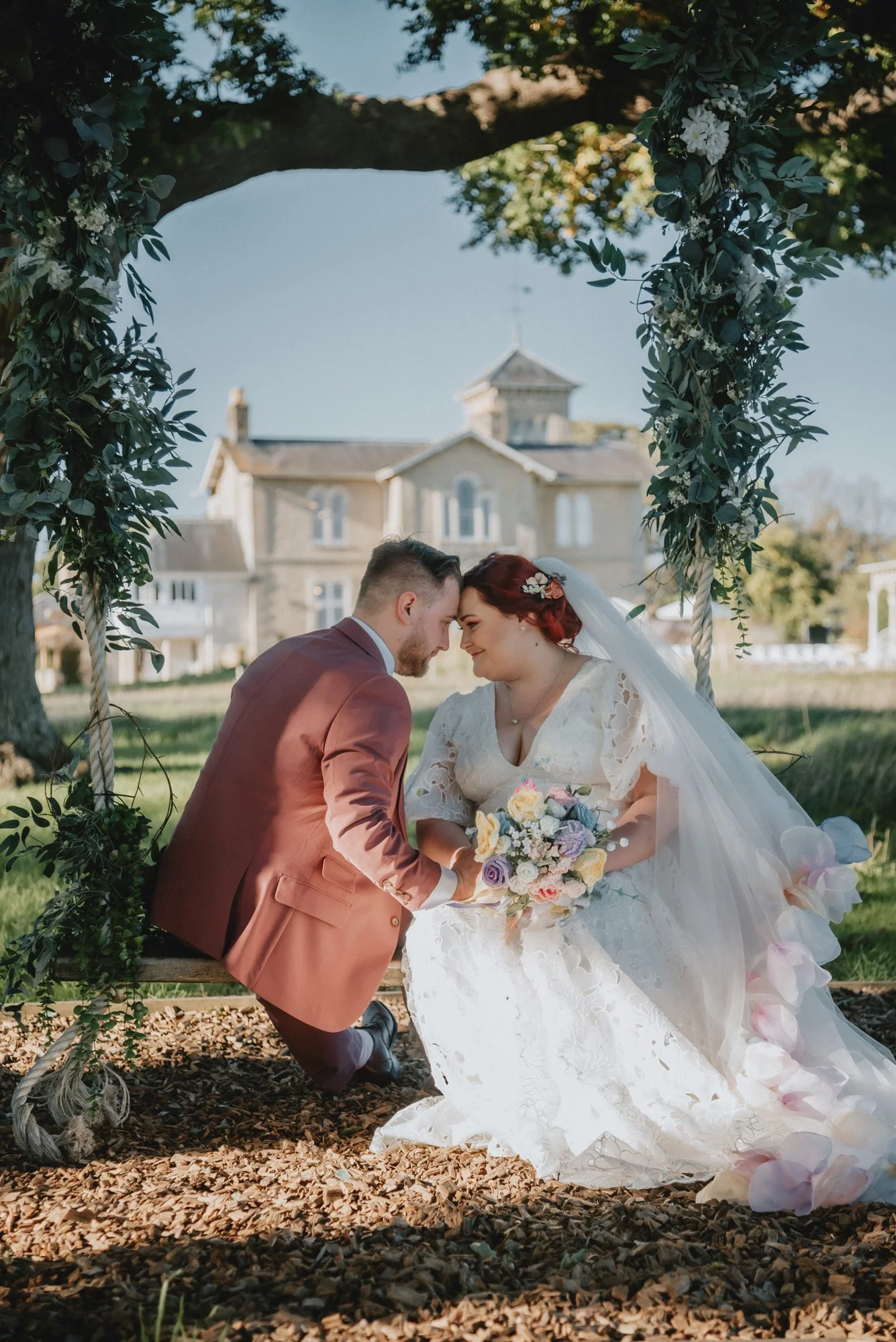 A bride and groom sitting face to face under a tree arch decorated with flowers, outdoors on a sunny day, with a large house in the background.