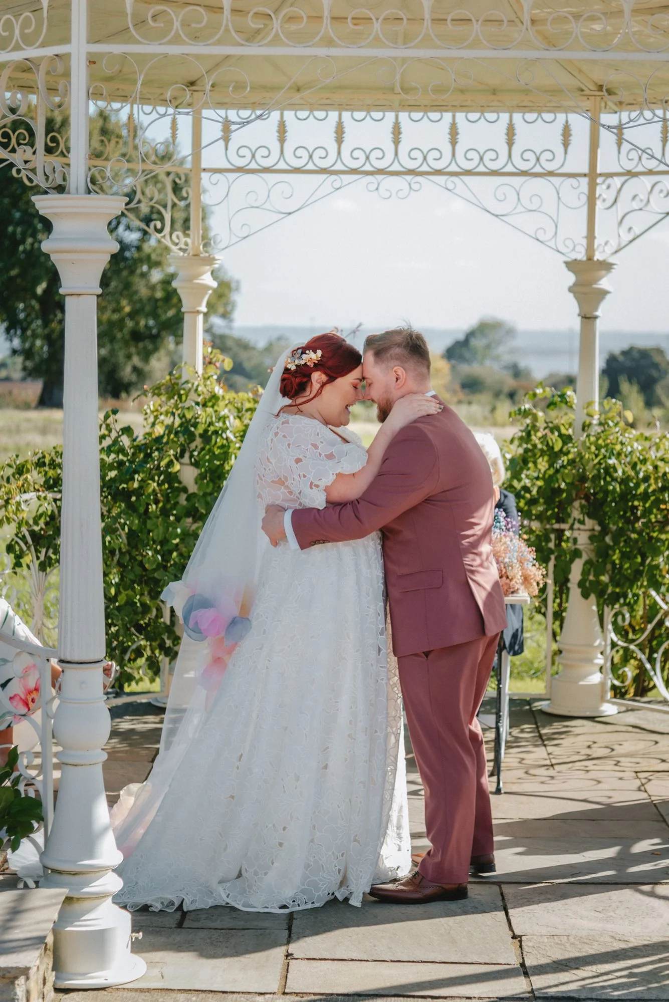 Bride and groom gently touching foreheads during their wedding ceremony outdoors, under a decorative gazebo with plants and trees in the background.