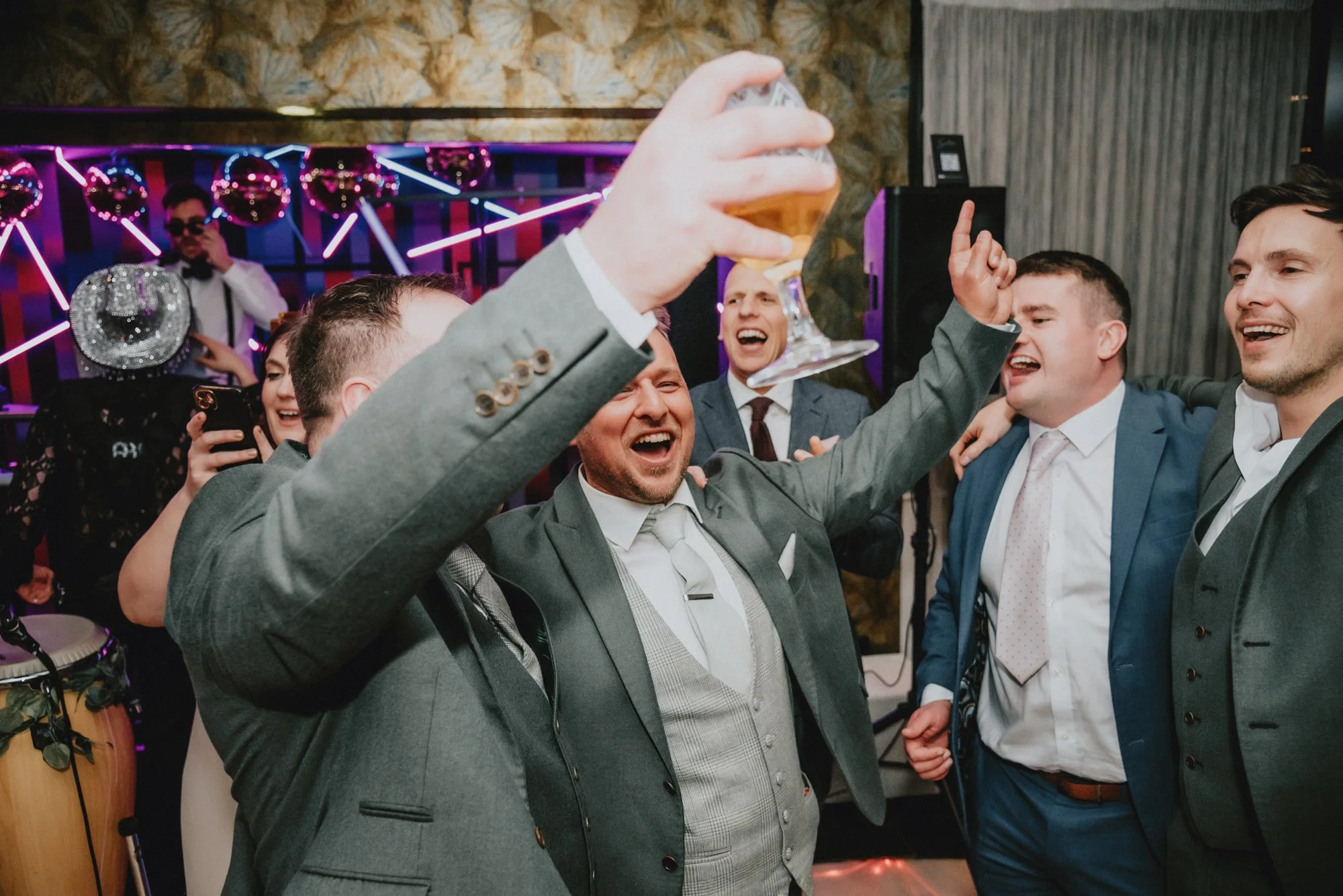 Group of men in suits celebrating and dancing at a party, with a man holding up a glass of beer in the foreground