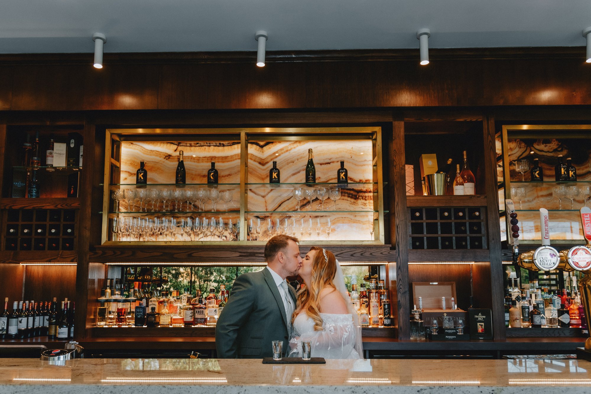 A man and woman kissing at a bar, with shelves of liquor bottles and glasses behind them.