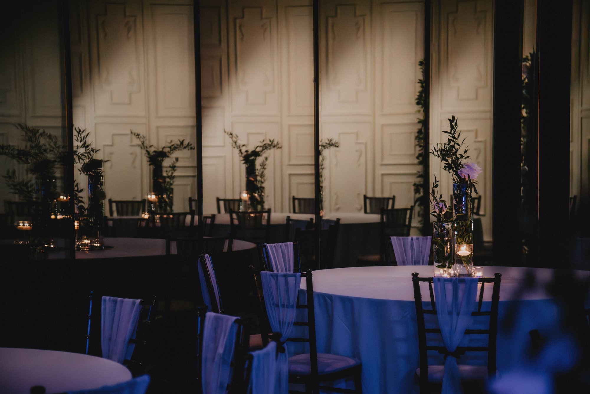 Elegant banquet hall with round tables draped in white tablecloths, decorated with floral centerpieces and candles, surrounded by chairs with sheer fabric and bows, illuminated softly with warm lighting.