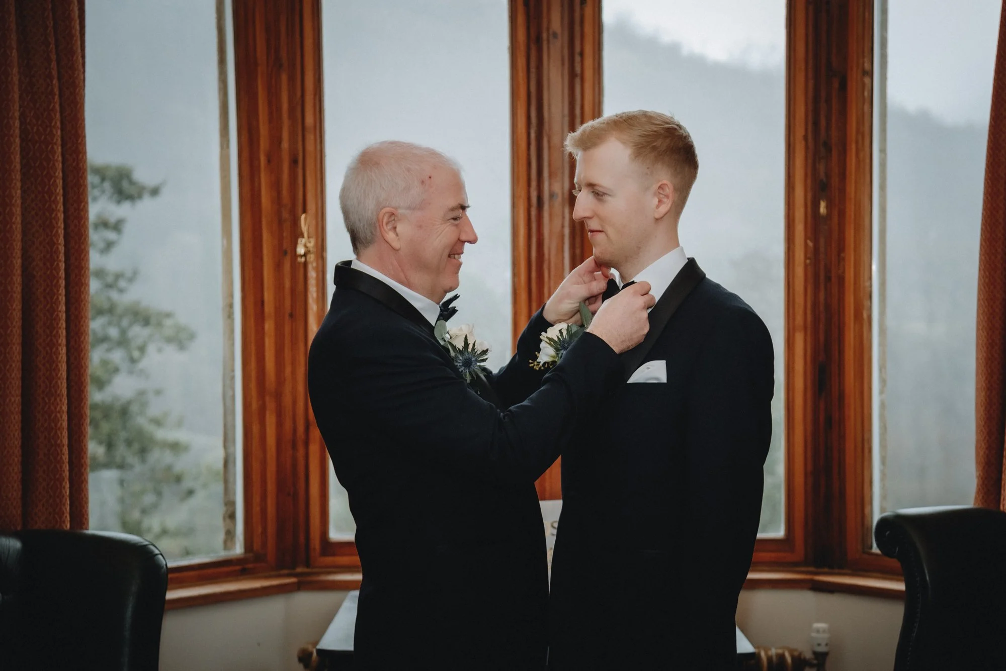 An older man helping a young man adjust his bow tie in a room with large windows behind them.