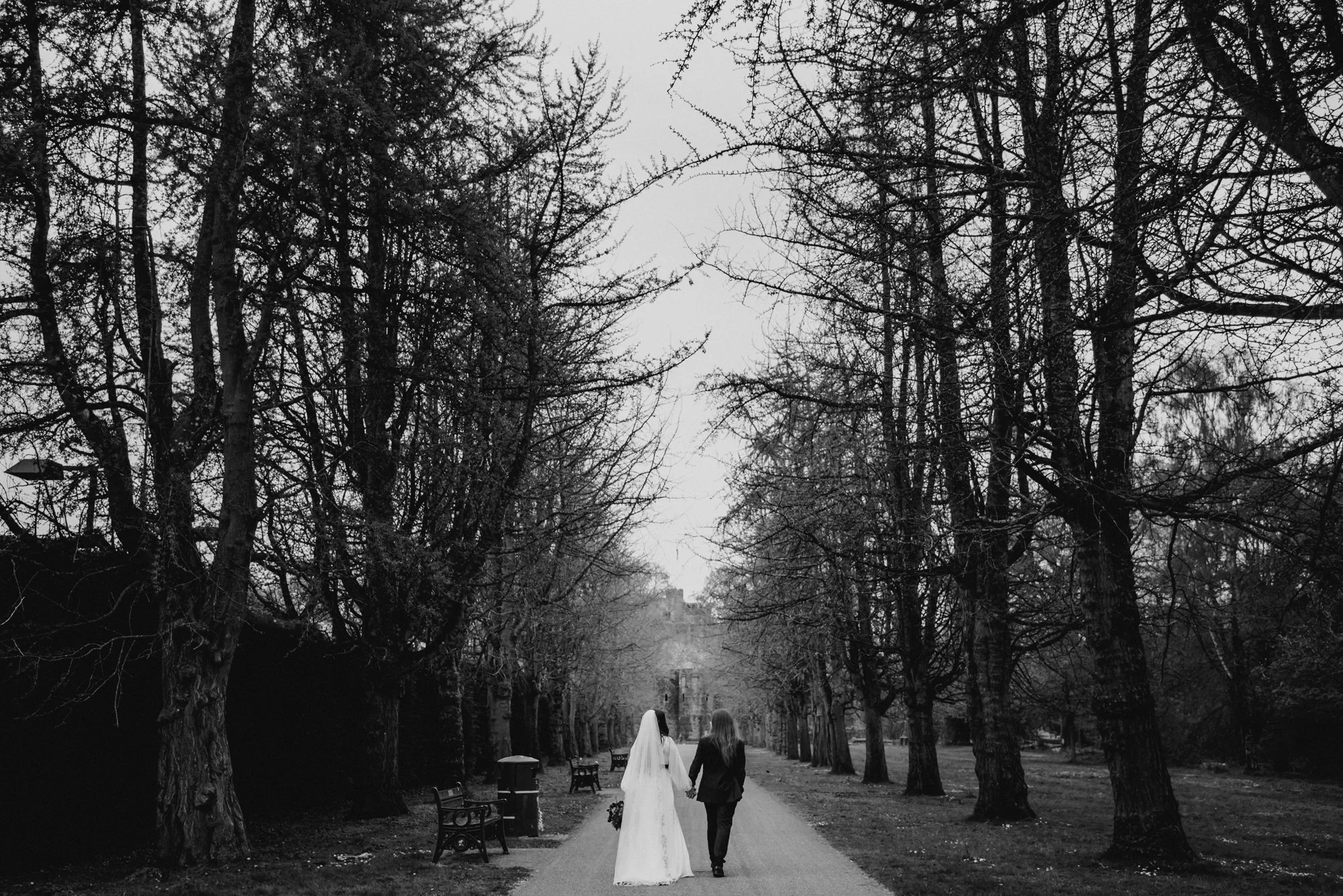 A bride and groom holding hands walking down a tree-lined path on a gray day