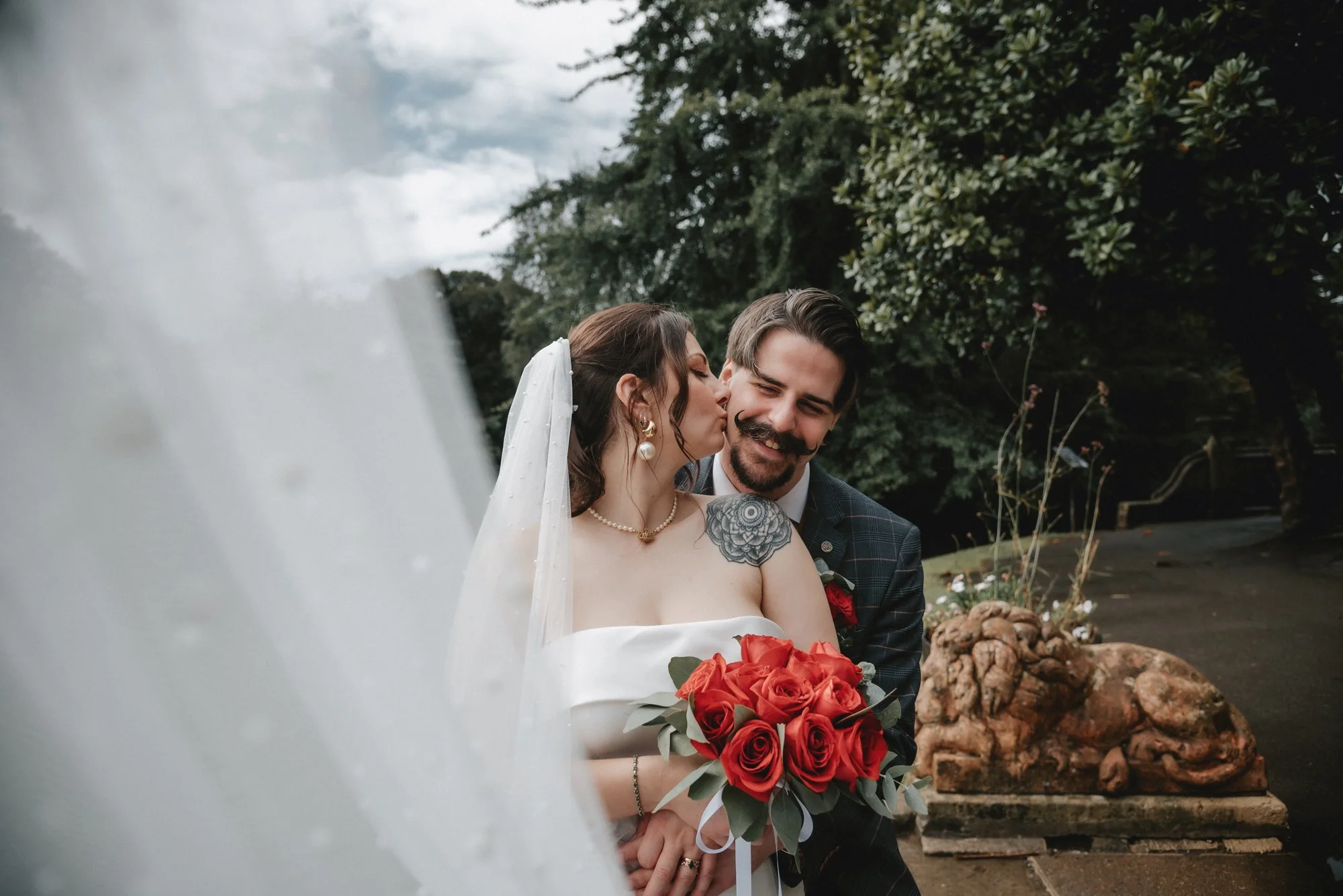 A bride and groom sharing a kiss outdoors, the bride holding a bouquet of red roses, with trees and a cloudy sky in the background.