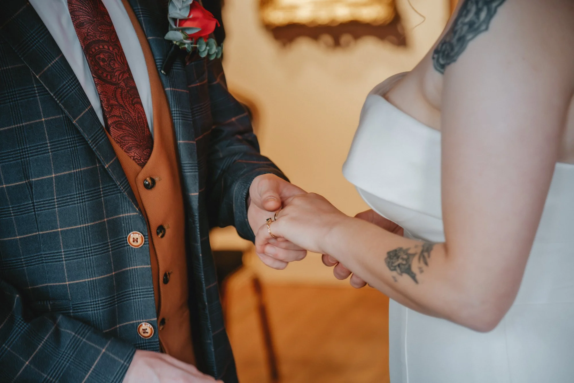 Close-up of a bride and groom holding hands during a wedding ceremony, with focus on their handshake and attire, including the groom's plaid suit and the bride's white dress with tattoos on her arm.