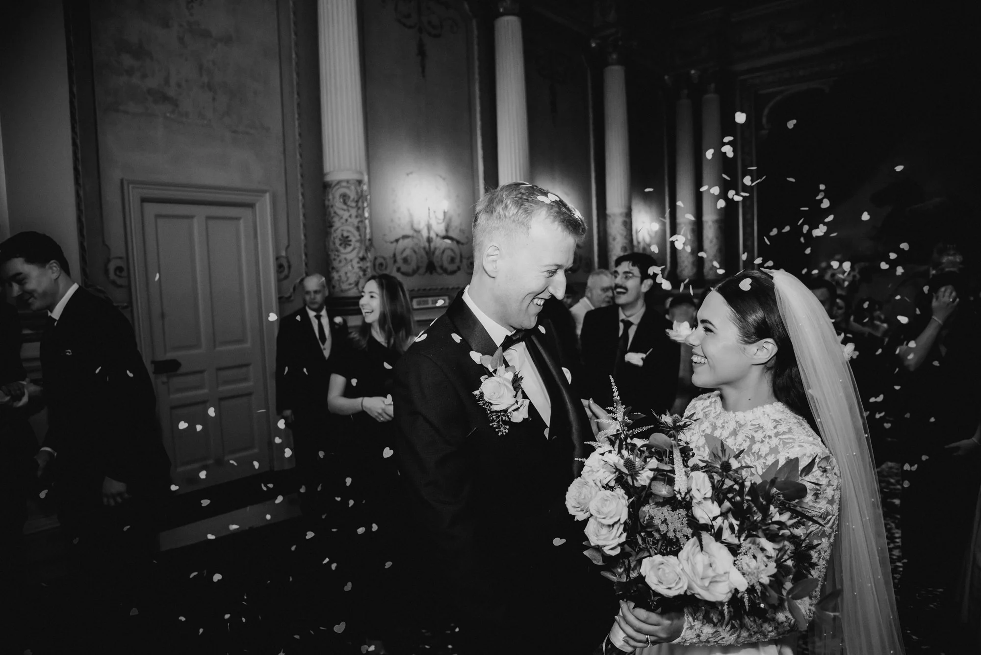 A black and white photo of a wedding reception, featuring a bride and groom smiling at each other, surrounded by guests, with confetti falling in the air.