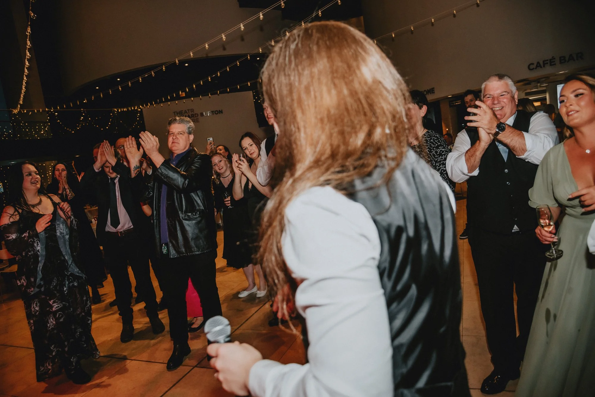 People at a celebration party dancing, clapping, and smiling while a woman with red hair, holding a microphone, stands in the foreground.