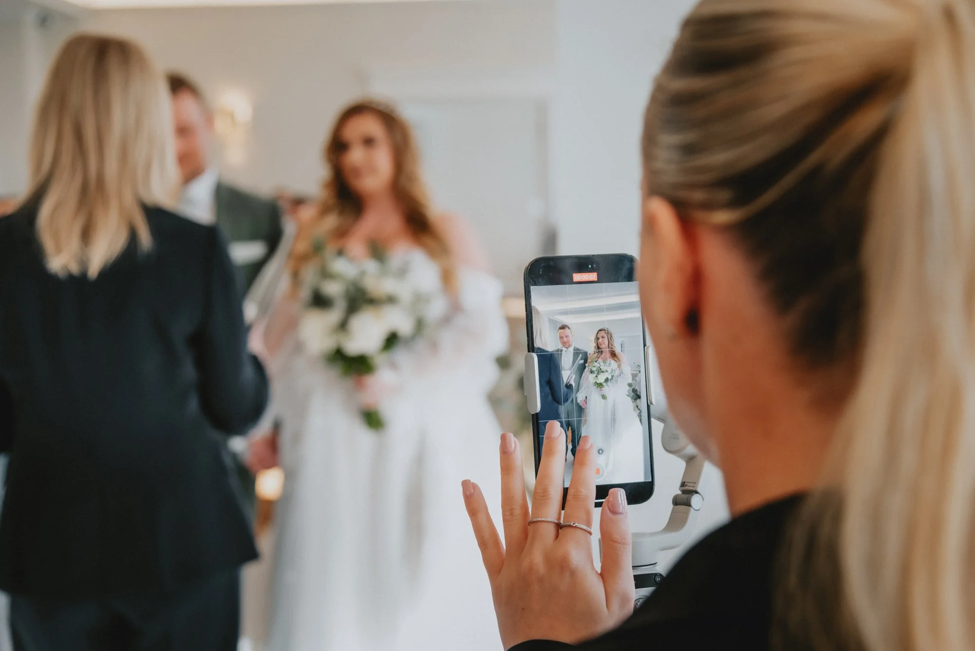 Person with blond hair taking a photo of a bride and groom with a smartphone during a wedding ceremony.