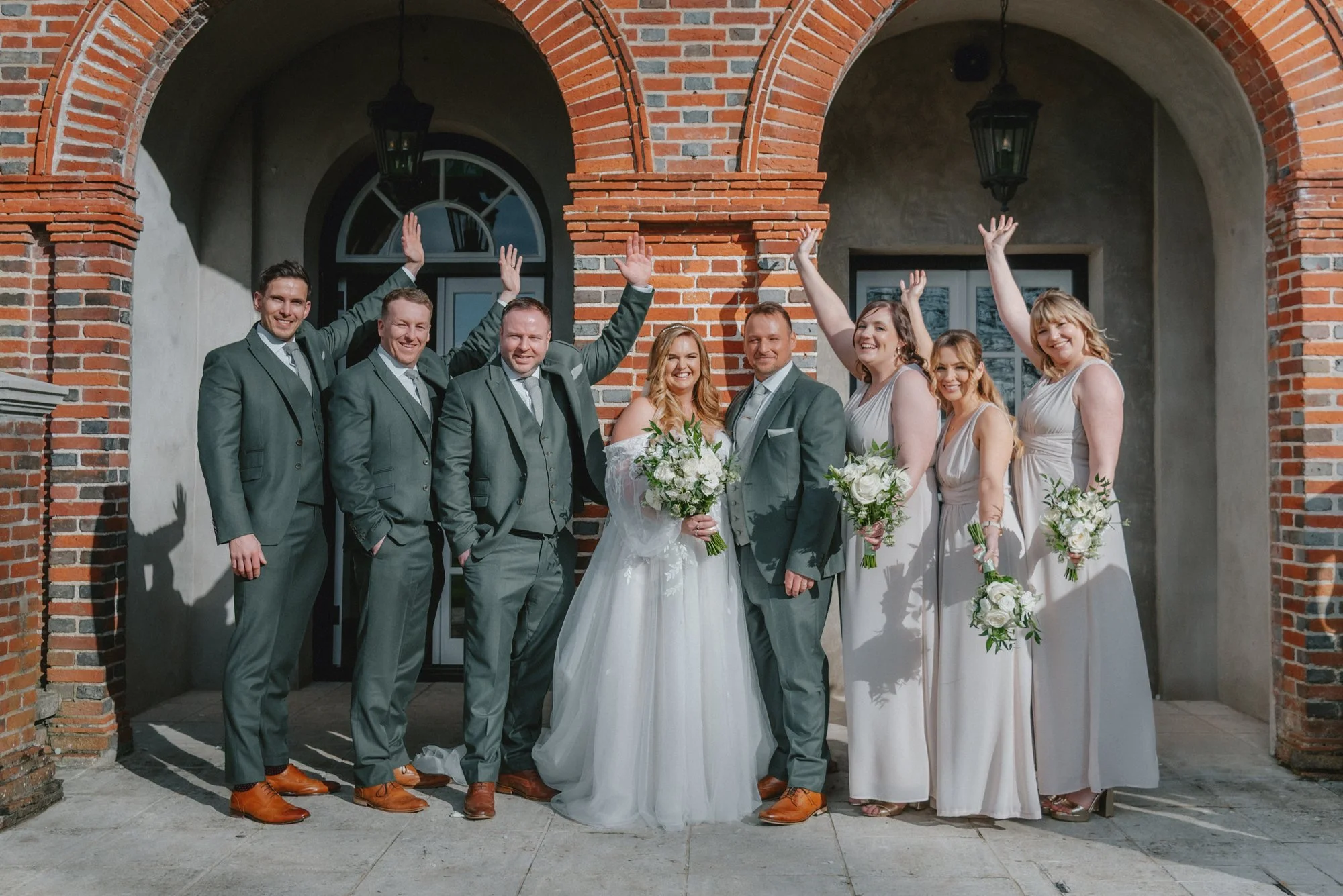 A group of wedding party members, including four groomsmen and four bridesmaids, posing in front of a brick building for a wedding photo. The bride and groom are in the center, smiling and holding bouquets. The bridesmaids and groomsmen have their ar
