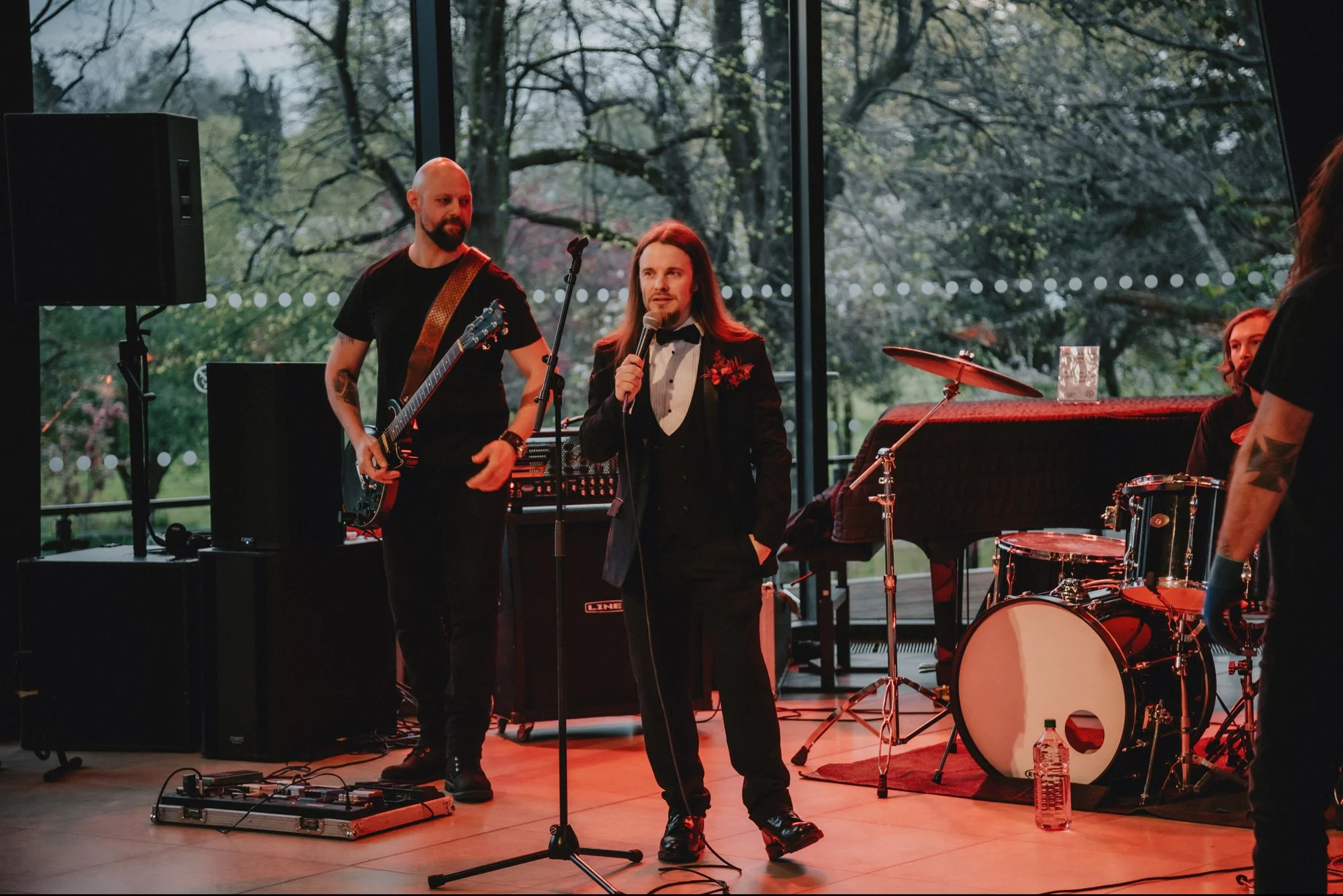 A band performing indoors with large windows showing trees outside. One person in a tuxedo-style suit with a microphone, two others with musical instruments including a guitar and a drum set.