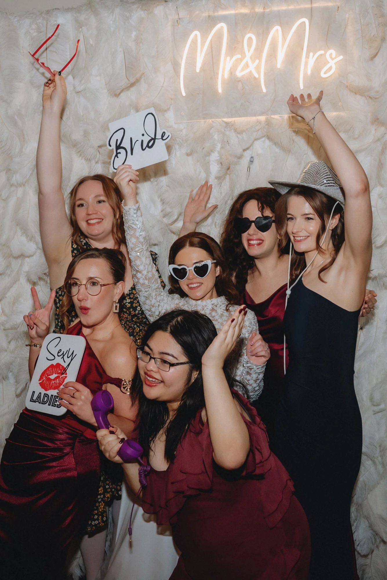 Group of six women celebrating at a bachelorette party with props, signs, and sunglasses in front of a white feathered backdrop and neon sign that reads "Mr & Mrs".