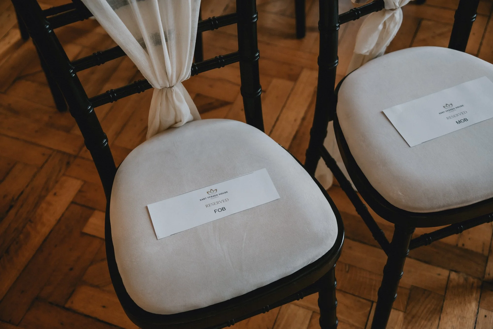 Two black chairs with white cushions and reserved signs on them, one for 'FOB' and one for 'MOB', placed on a hardwood floor. The chairs have white fabric tied to the back.