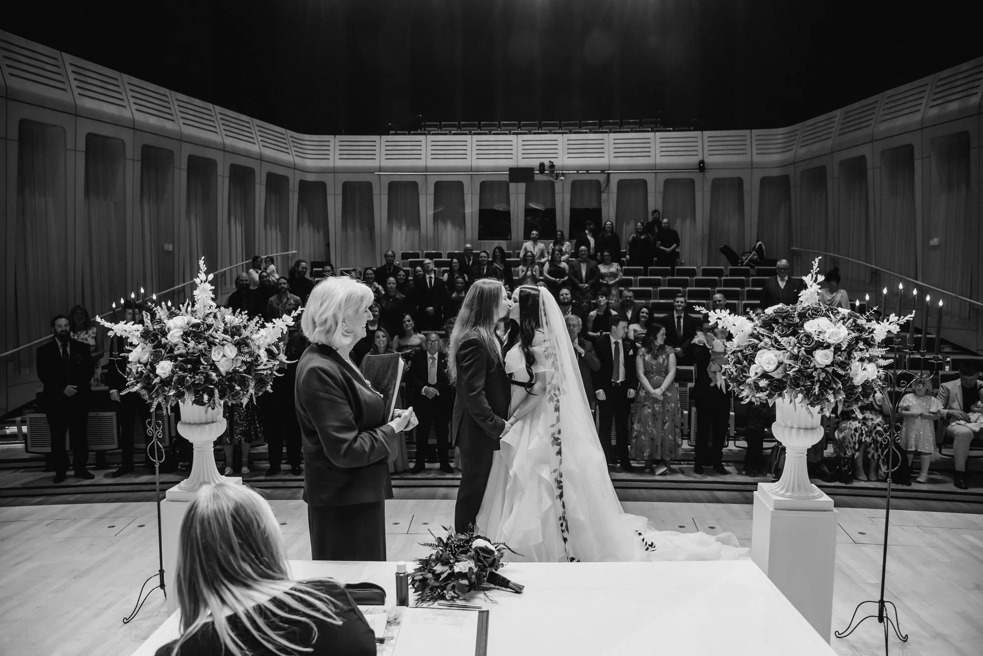 A black and white photo of a wedding ceremony with two brides kissing, an officiant standing nearby, floral arrangements, and an audience in the background in a modern auditorium.