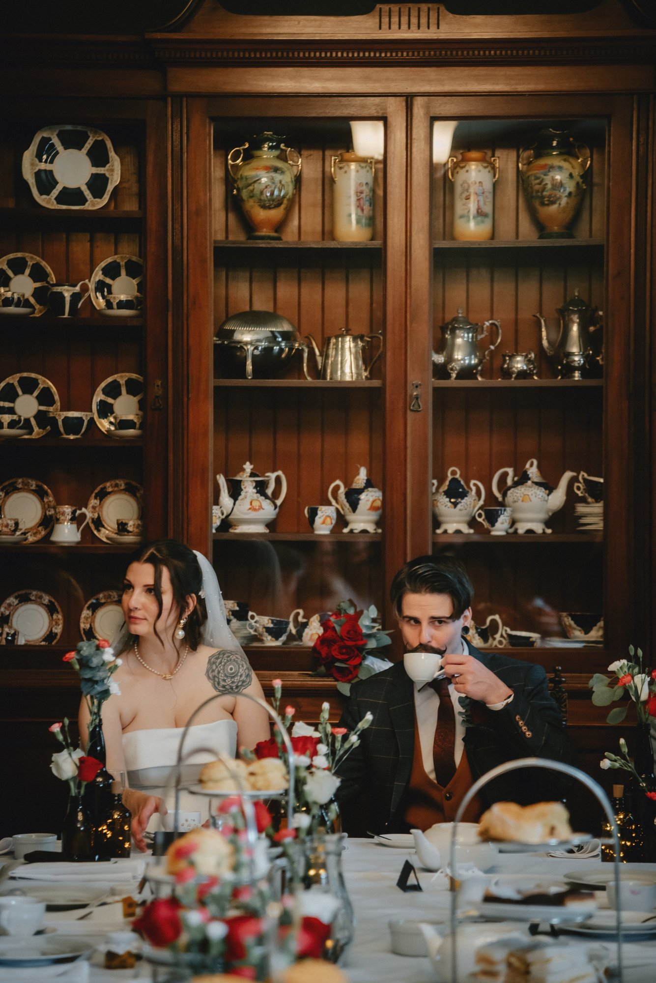 A bride and groom sit at a decorated table during a wedding reception, with the bride wearing a strapless white dress and veil, and the groom wearing a dark suit and drinking from a teacup, in a room with wooden cabinet displaying china.