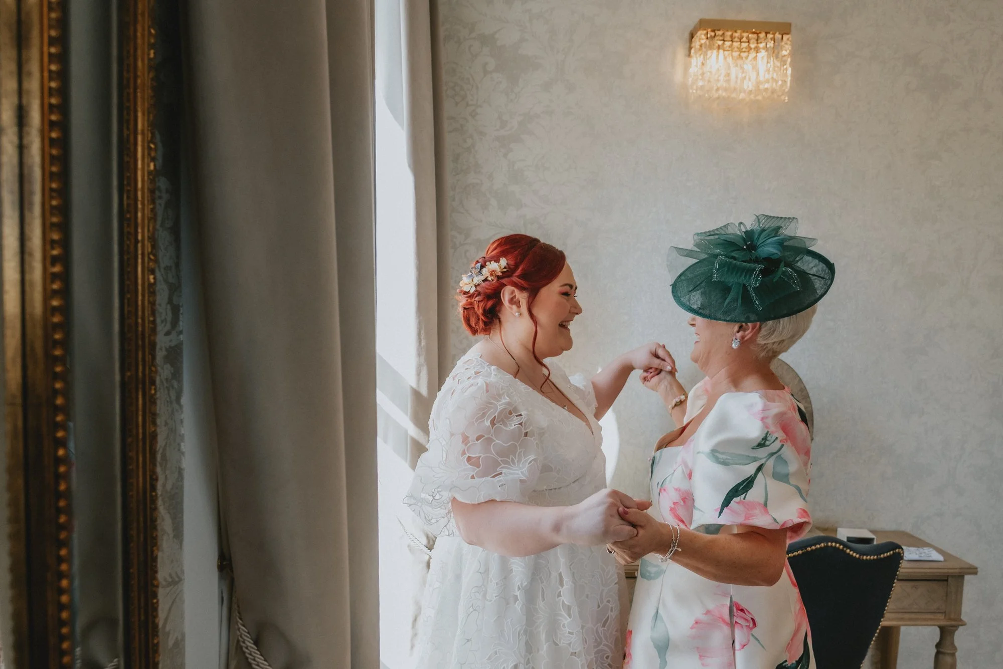 A bride and an older woman sharing a joyful moment, holding hands and smiling at each other in a well-lit room with elegant decor.