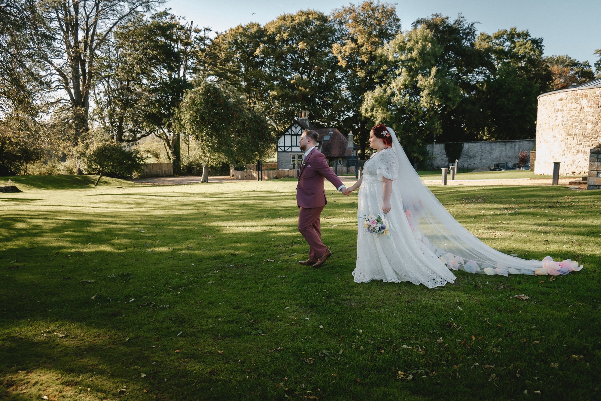 A bride and groom holding hands outdoors on a sunny day in a grassy park, with trees and old stone buildings in the background. The bride is wearing a white lace wedding dress with a long train and a veil, holding a bouquet, and the groom is dressed 
