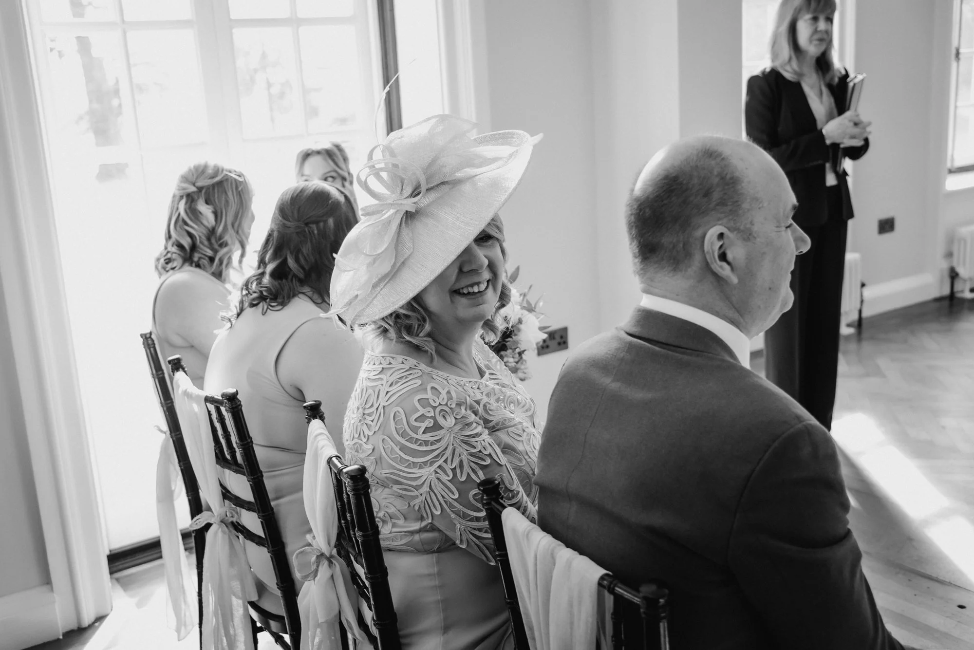 Black and white photo of a woman wearing a large hat with decorative elements, smiling and sitting beside a man in a suit. Several women with styled hair are seated nearby, and a woman in the background is standing with a notepad.