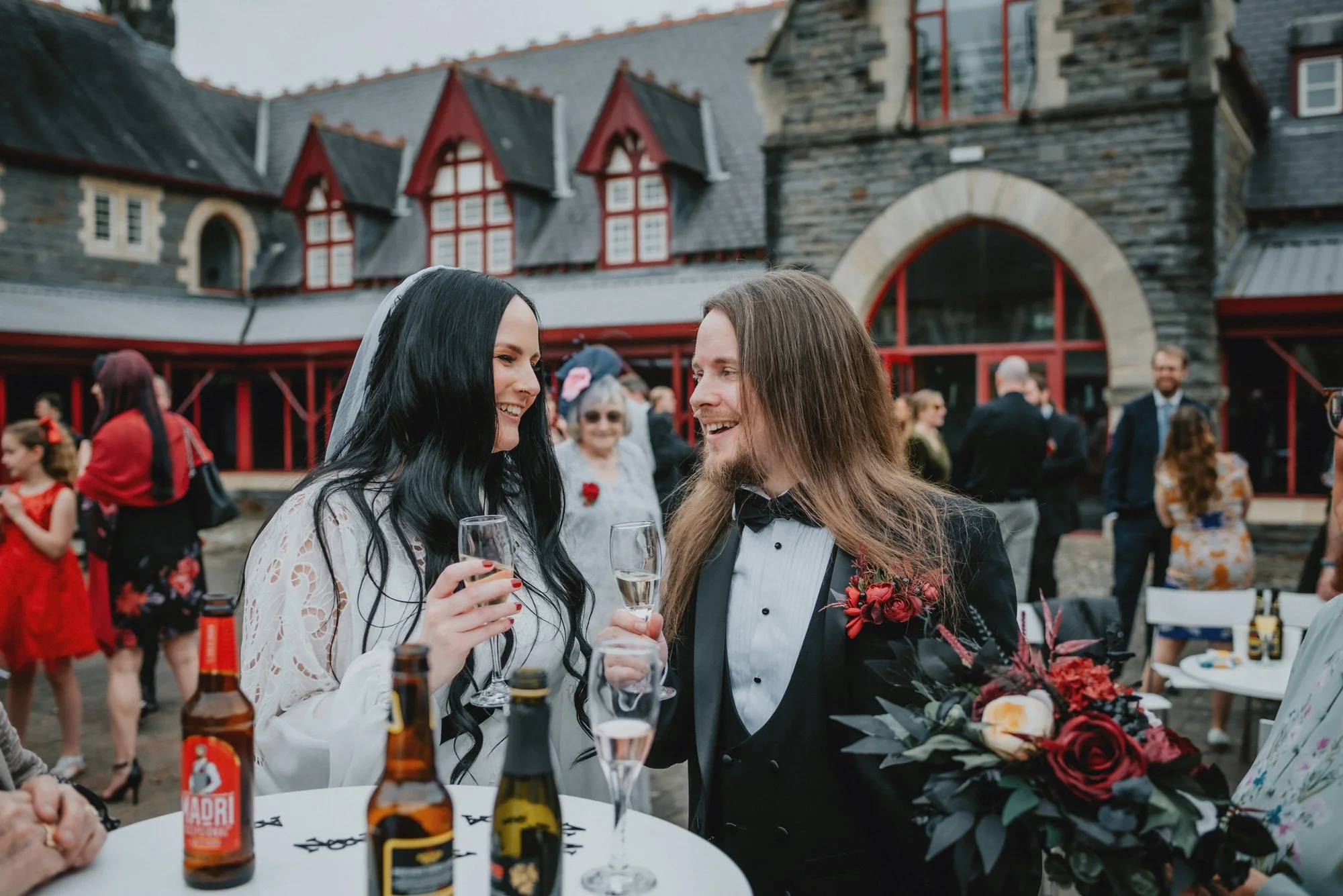 A bride and groom smiling and holding glasses of champagne at their wedding reception outside a large stone and timber building, surrounded by guests.