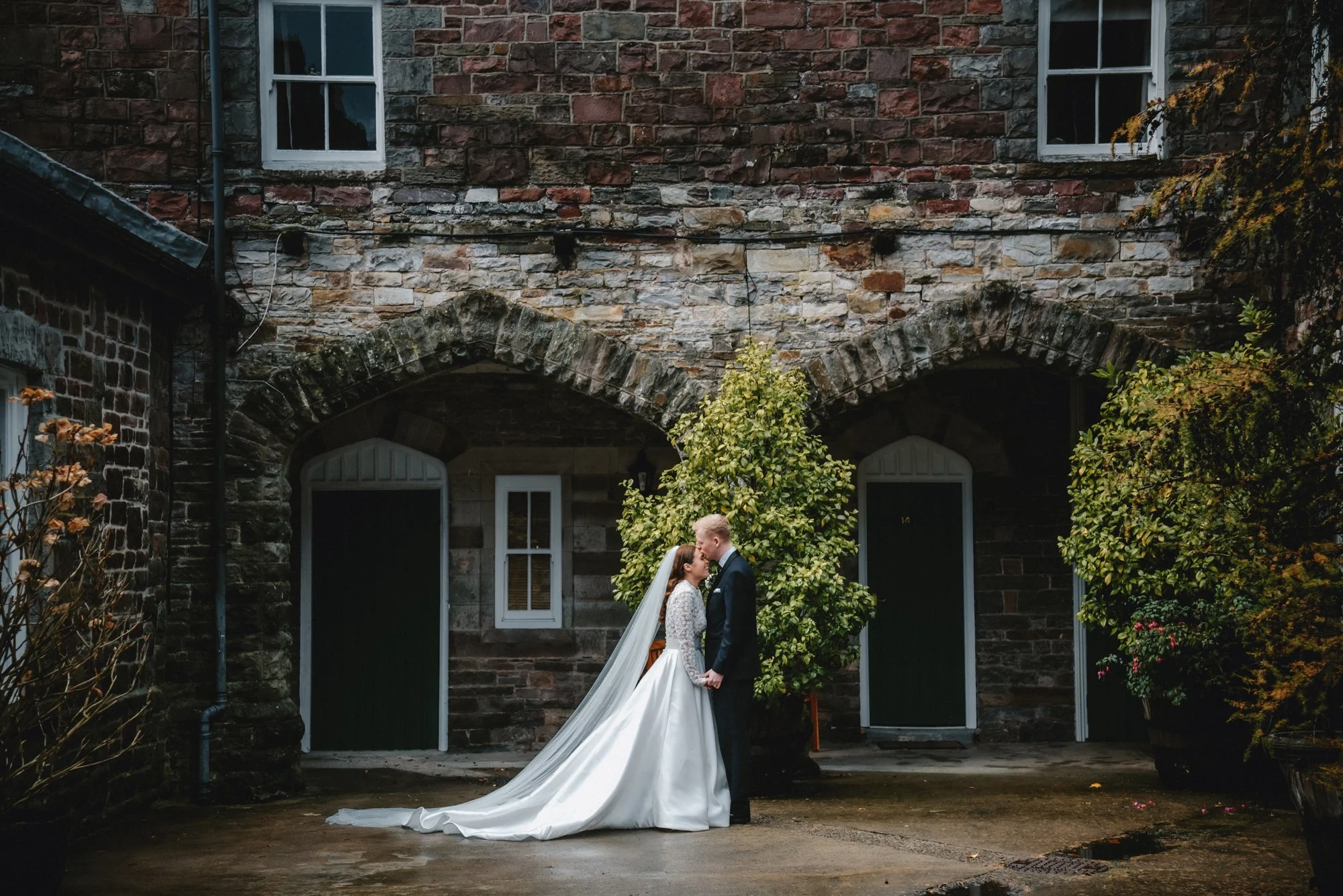 A bride and groom stand holding hands and kissing in front of a brick building with two arched doorways and windows, surrounded by plants and greenery.