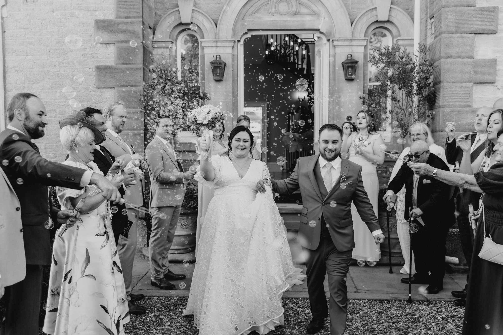 Black and white photo of a bride and groom walking out of a building, surrounded by guests blowing bubbles and celebrating on a wedding day.