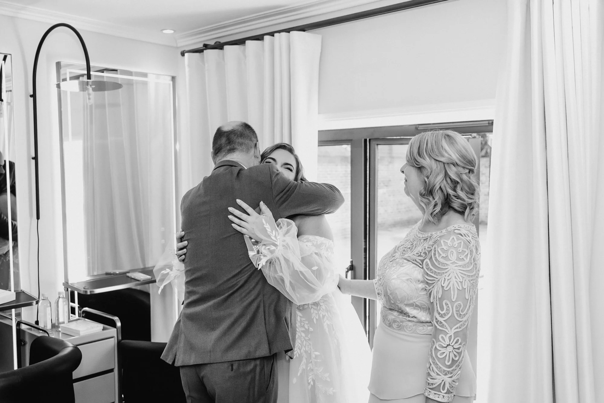 A bride being hugged by a man, with a woman watching, inside a room with curtains and a window.