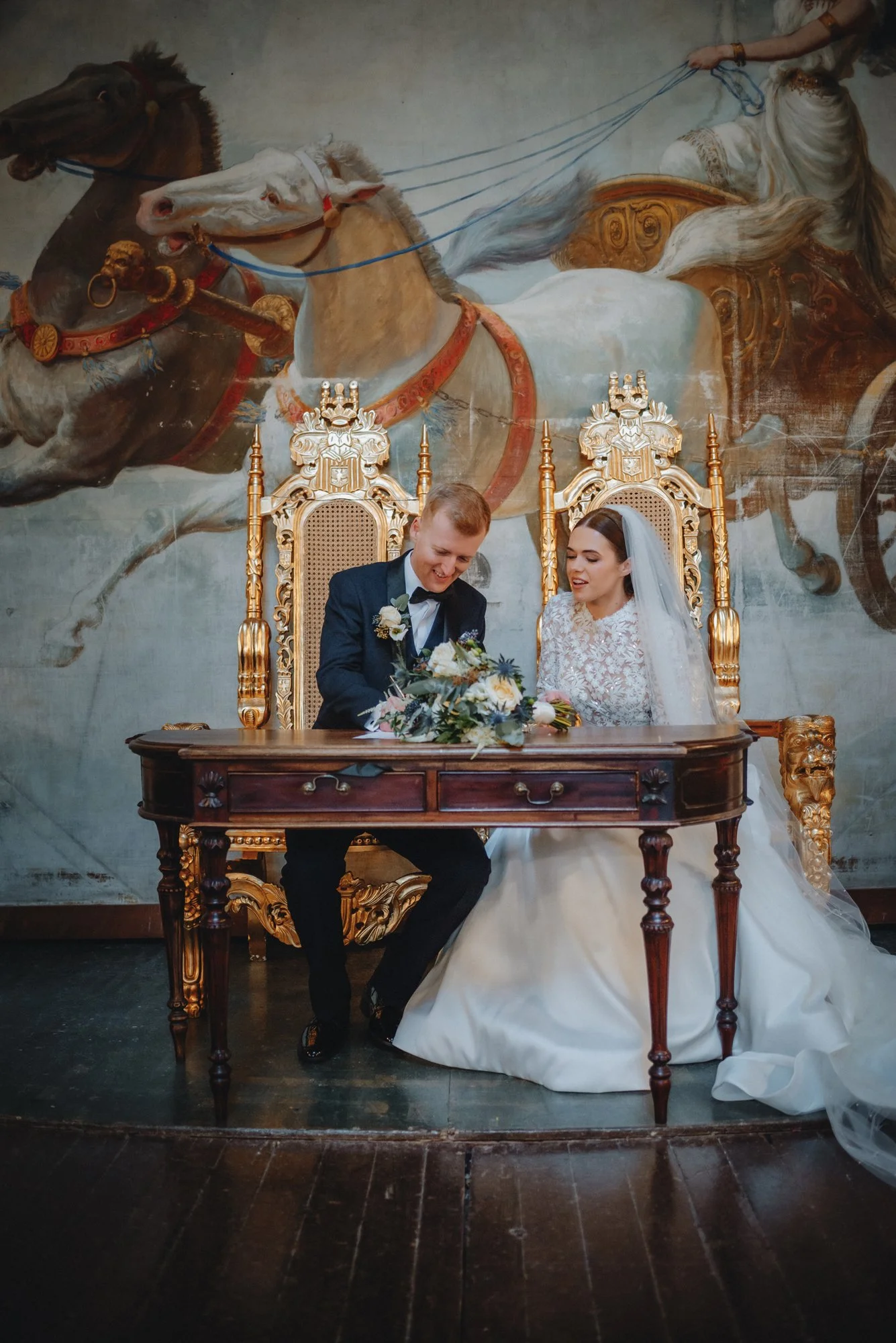 A bride and groom signing a marriage document at a wedding ceremony, seated at a dark wooden table, with a backdrop of a mural depicting a chariot pulled by horses.