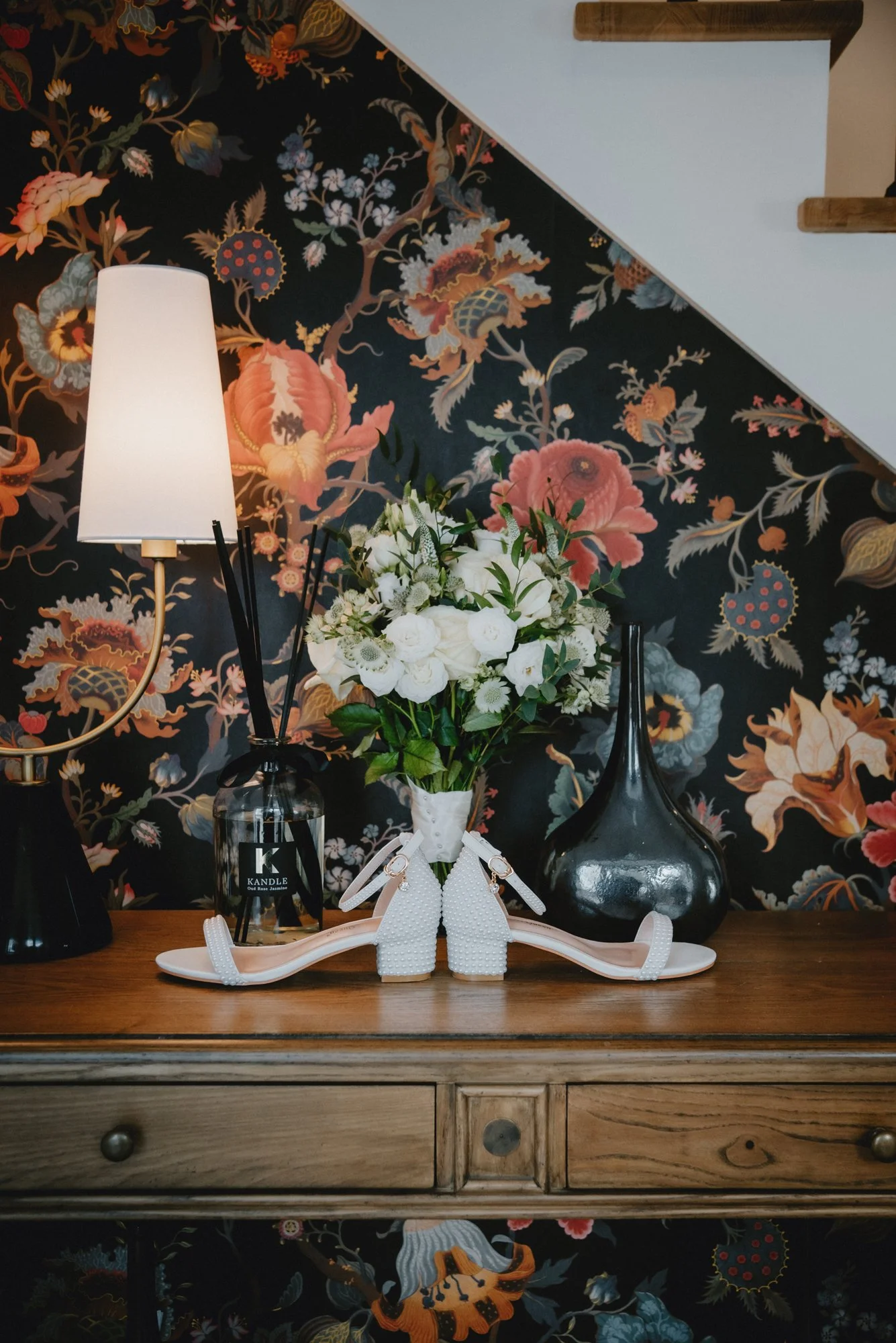 A floral arrangement of white flowers in a white vase, placed on a wooden table with white heeled shoes in front, and a black vases, a table lamp, and a diffuser on either side, against a floral patterned wallpaper.