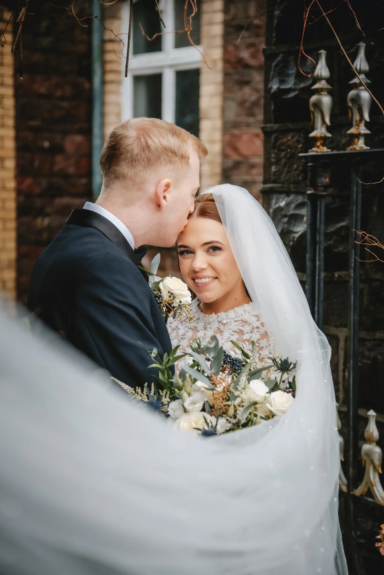 A bride and groom sharing a tender moment on their wedding day outdoors. The groom is kissing the bride's forehead, and she is smiling warmly. The bride is wearing a lace wedding dress and veil, holding a bouquet of white flowers, while the groom is 