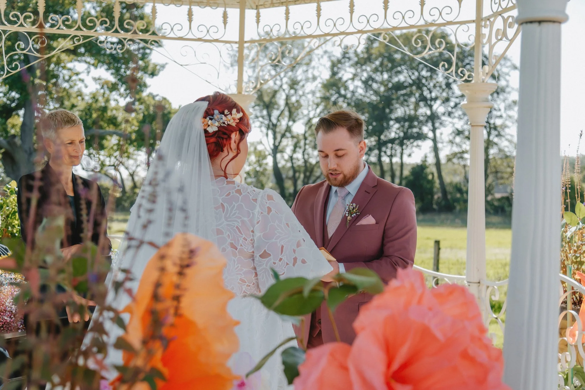 A wedding ceremony outdoors under a white arbor, with a bride and groom exchanging vows. The bride has red hair and wears a white lace dress and veil, adorned with small flowers. The groom has short hair and a beard, dressed in a maroon suit with a t
