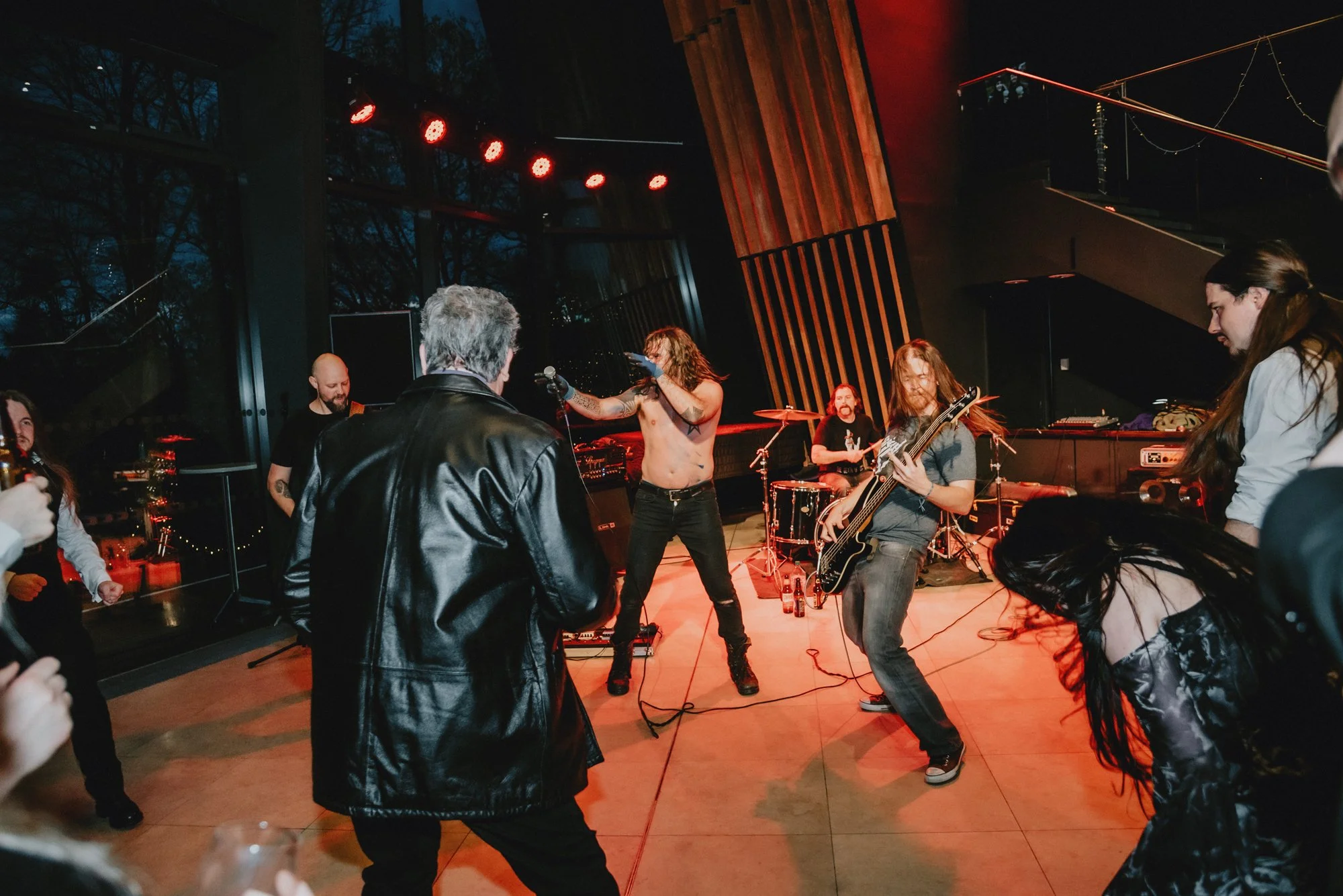A live music performance with a band playing on stage, singer without shirt, others playing guitar and drums, audience dancing nearby, warm red stage lighting, indoor setting with large windows.