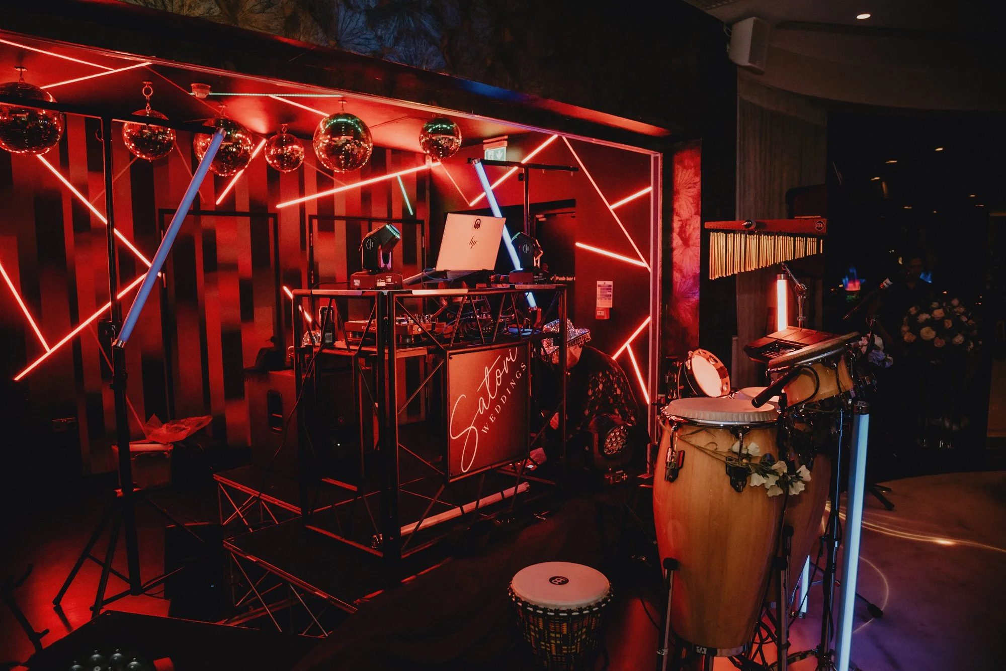 A DJ set on a stage at a wedding reception with red neon lighting, disco balls, musical instruments including drums and a xylophone, and a sign that reads 'Saton Weddings'.