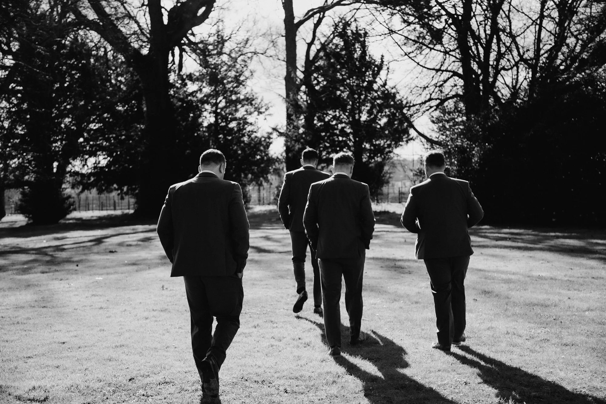 Five men in suits walk away on a grassy area with trees in the background, black and white photo